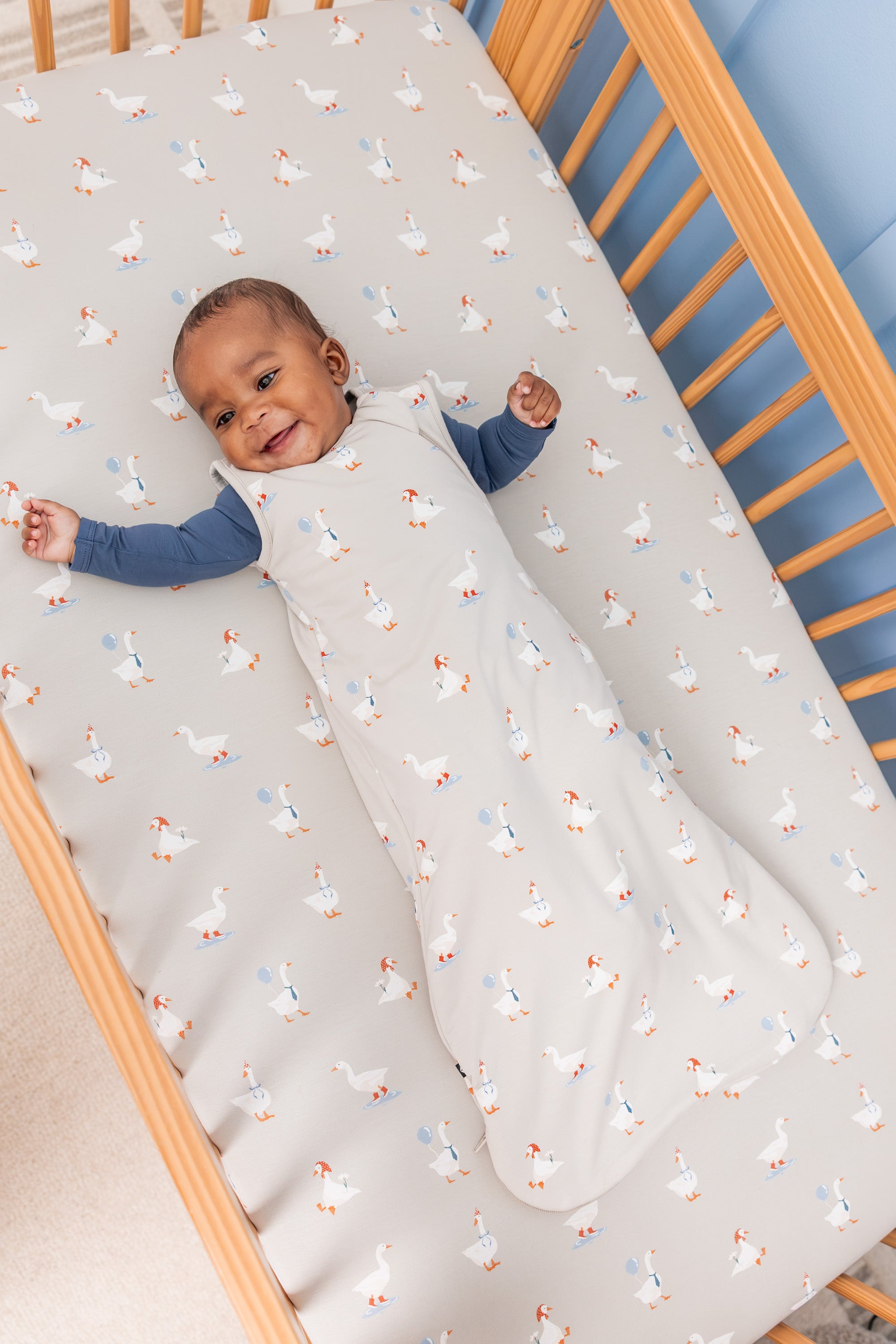 Baby wearing Sleep Bag in Oat Silly Goose 1.0 print over oxford zippered romper lying in a crib over oat silly goose print crib sheet