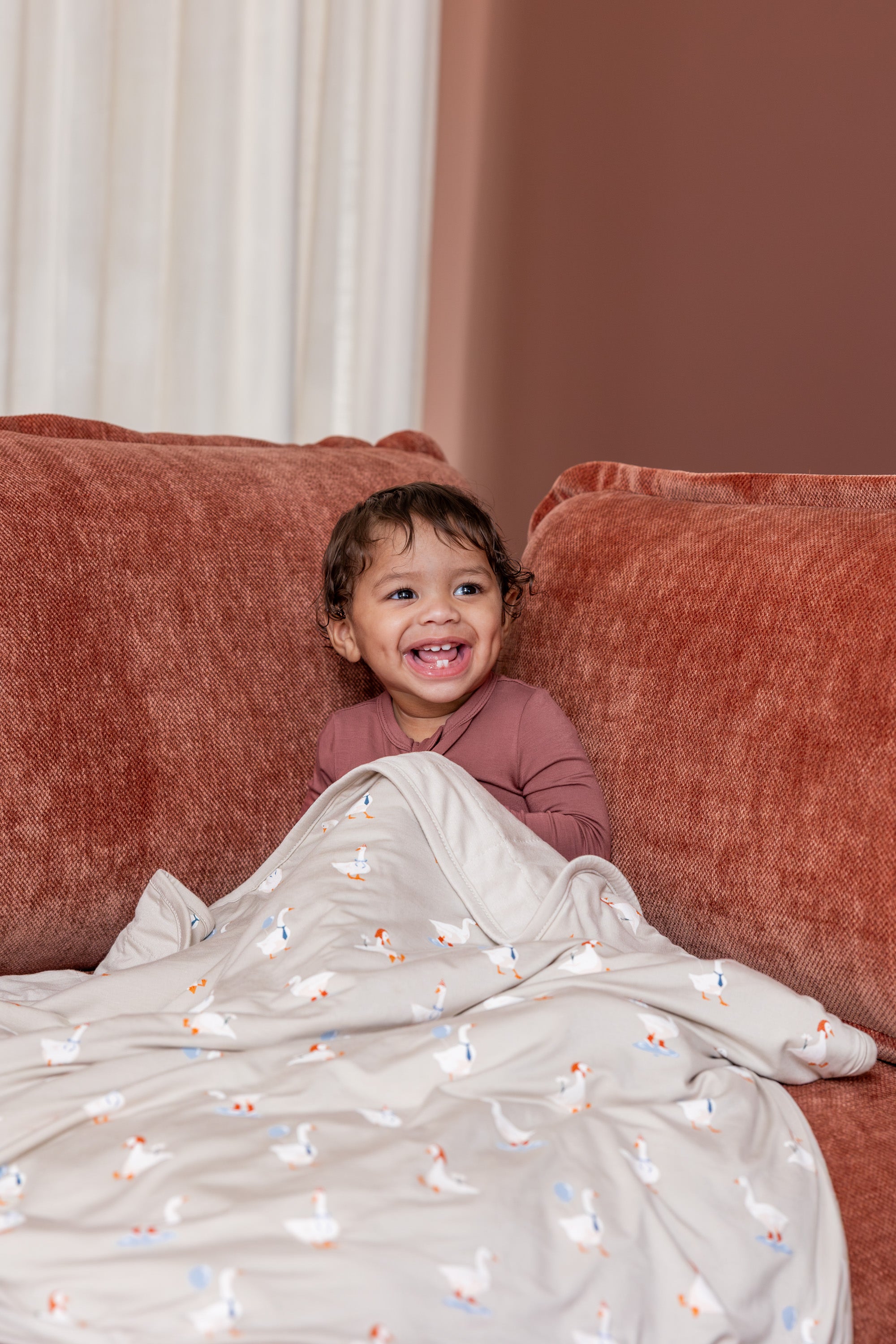 Baby sitting with Baby Blanket in Oat Silly Goose 1.0 print on a brown sofa wearing ember color zippered romper