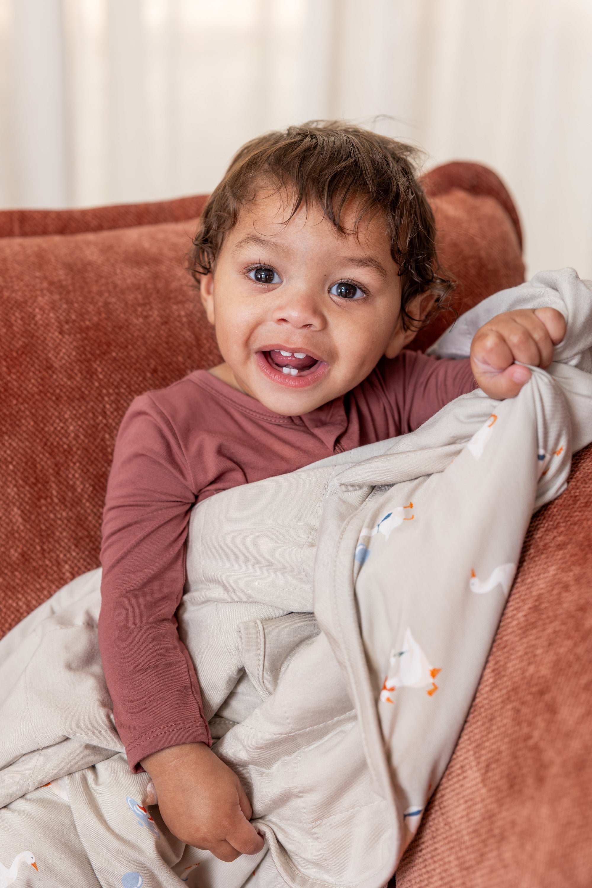 Baby sitting with Baby Blanket in Oat Silly Goose 1.0 print on a brown sofa wearing ember color zippered romper
