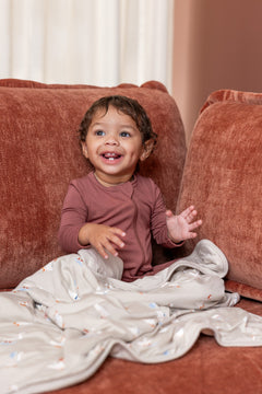 Baby sitting with Baby Blanket in Oat Silly Goose 1.0 print on a brown sofa wearing ember color zippered romper