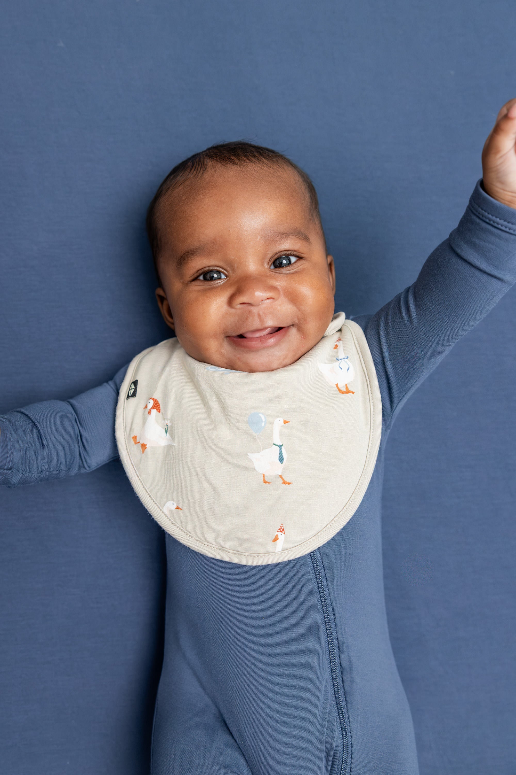 Baby wearing Cushy Bib in Oat Silly Goose print on oxford color zippered romper lying on blue sheet