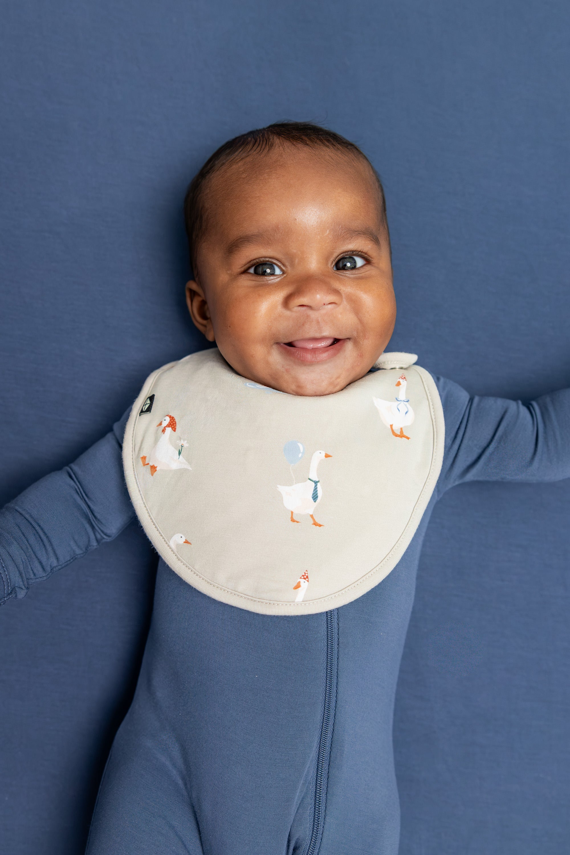 Baby wearing Cushy Bib in Oat Silly Goose print on oxford color zippered romper lying on blue sheet