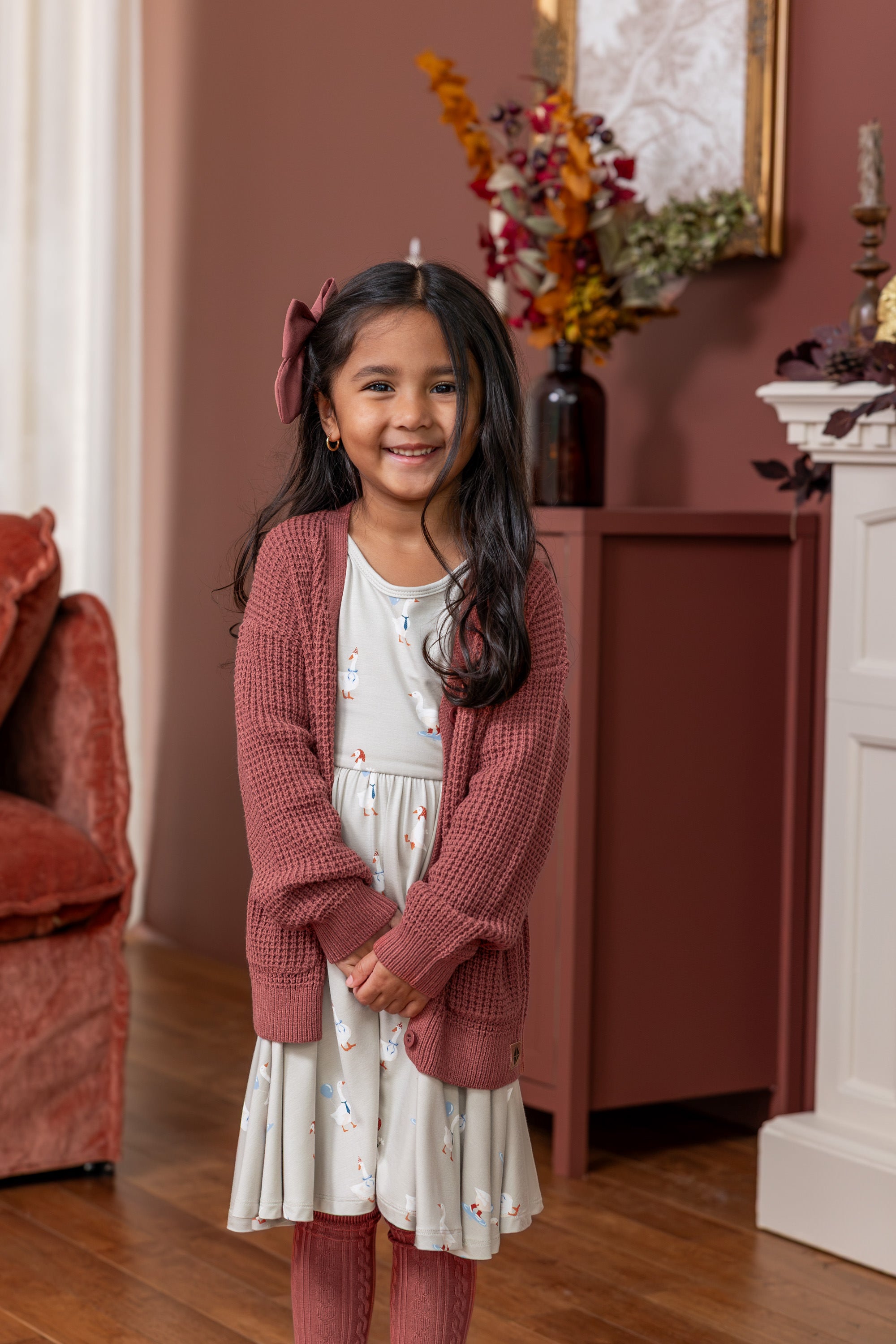 Toddler wearing Long Sleeve Twirl Dress in Oat Silly Goose print and ember color chunky knit waffle cardigan with ember color knee high socks standing in a living room 
