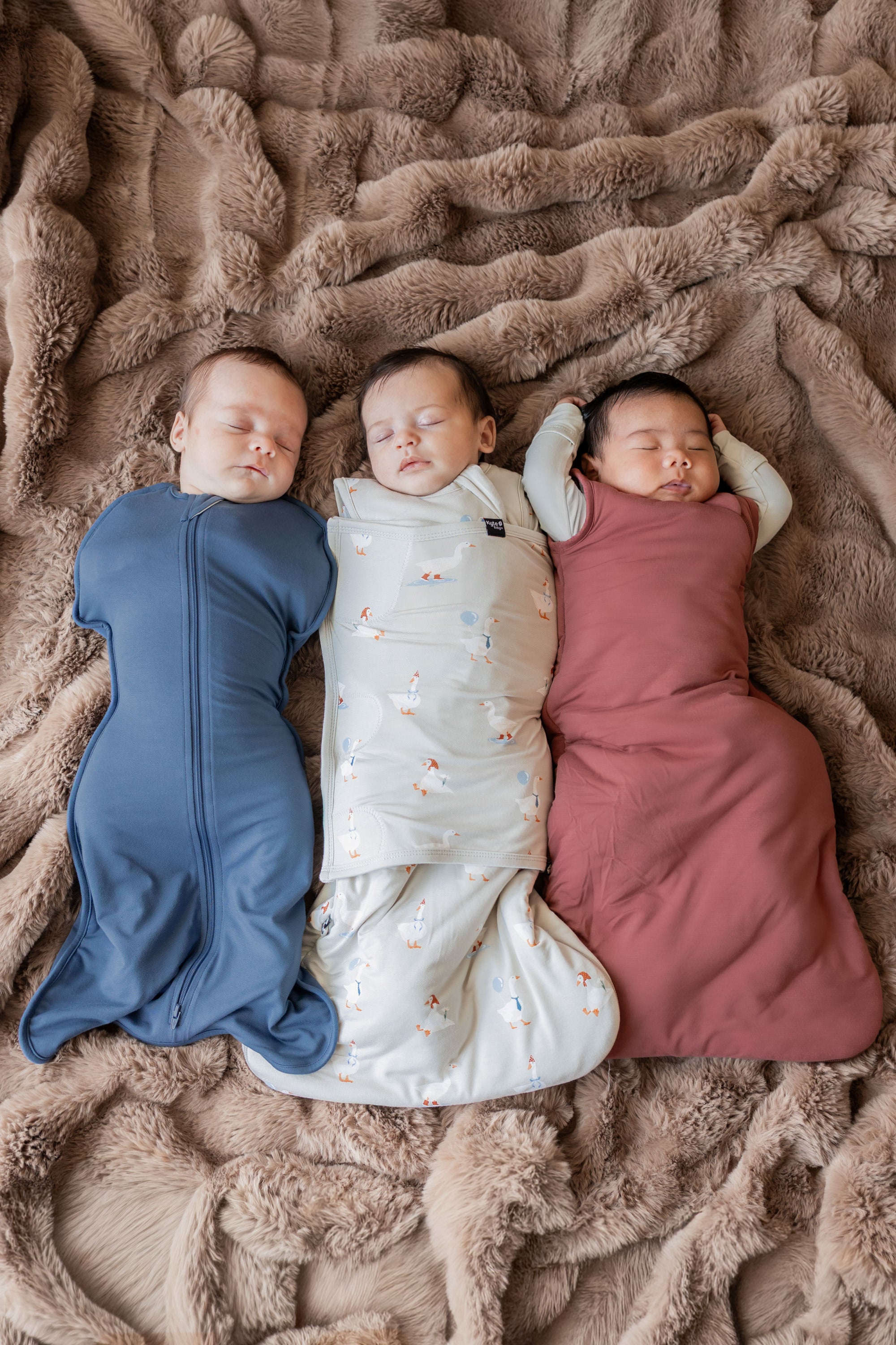 Three babies sleeping together wearing Sleep Bag Swaddler in Oat Silly Goose 1.0 print, Swaddle bag in oxford color and Ember color sleep bag  over brown blanket 