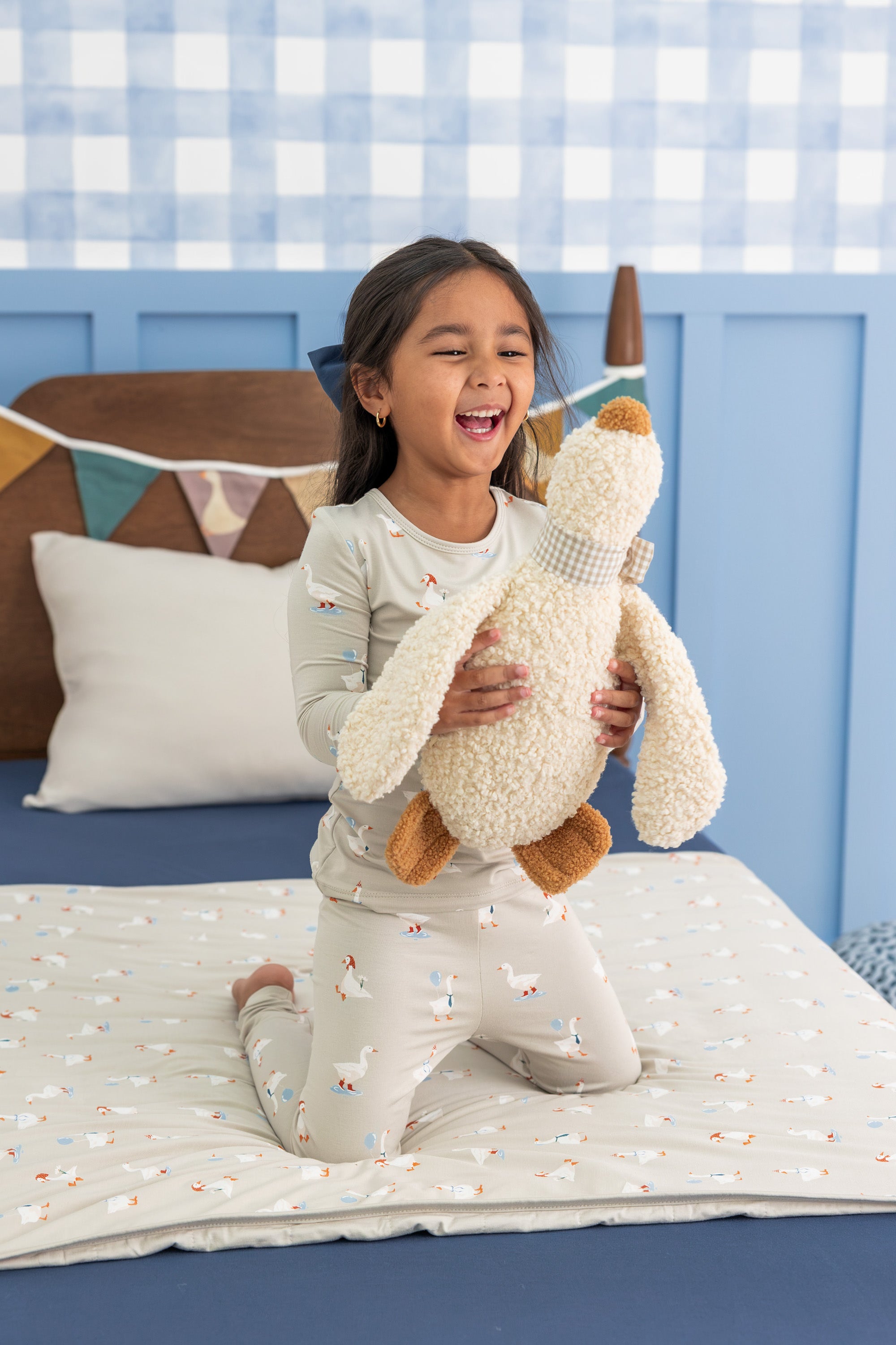Girl holding an oat color silly goose stuff toy wearing Long Sleeve Pajamas in Oat Silly Goose print sitting on a bed