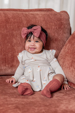 Baby wearing Long Sleeve Twirl Bodysuit Dress in Oat Silly Goose print, with ember knotted bow head band and knee high socks on a brown sofa