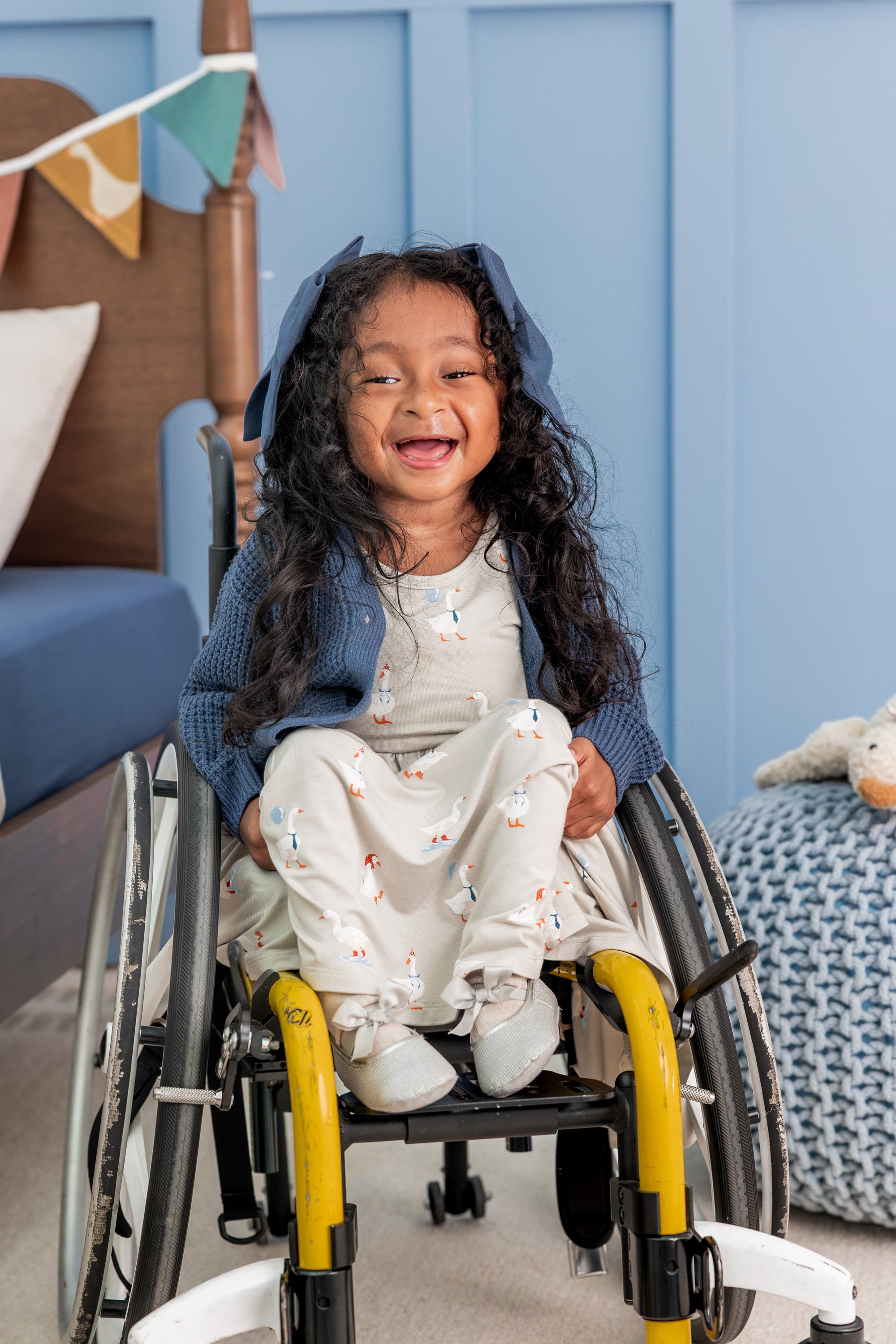 Girl wearing Twirl Dress in Oat Silly Goose print with chunky knit waffle cardigan in oxford color sitting on a wheel chair beside a bed
