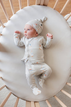 Baby wearing Take Me Home Set with Cap in Oat Silly Goose print lying in a crib over oat color sheet