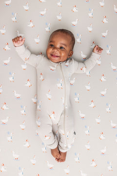 Baby wearing Zippered Romper in Oat Silly Goose print over similar print sheets