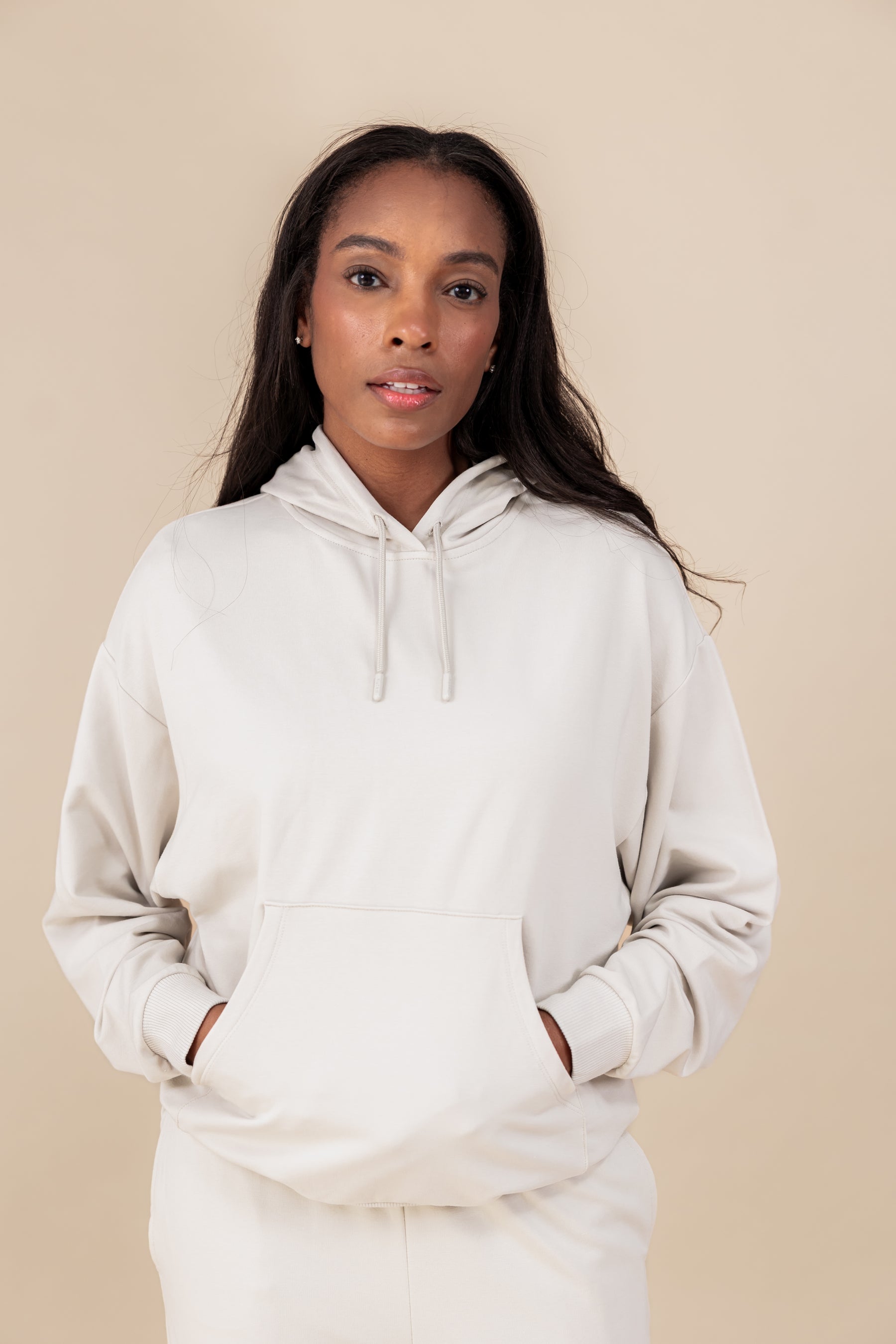 Modal wearing Women's Hoodie in Oat color with her hands in the pockets over similar color jogger pants on a beige background