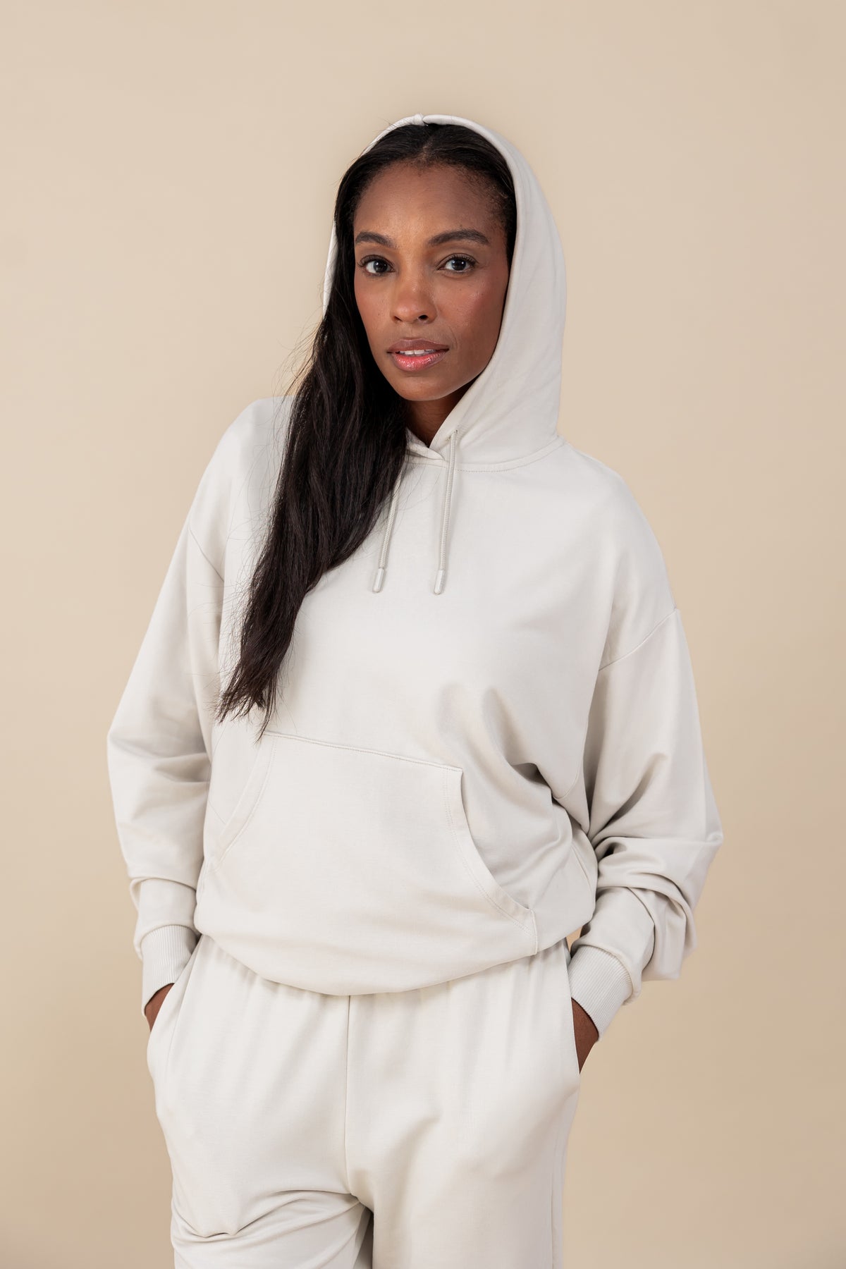 Modal wearing Women's Hoodie in Oat color over similar color jogger pants on a beige background