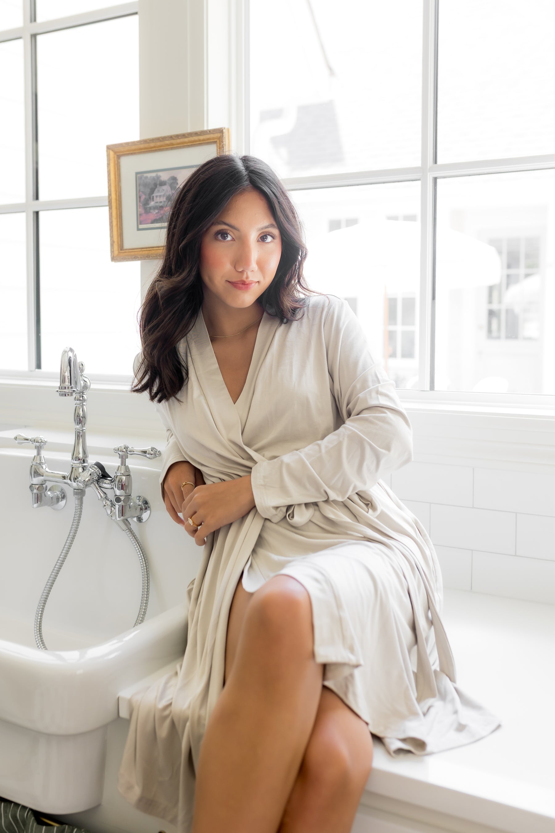 Female sitting on a white counter beside a sink wearing the Women’s Lounge Robe in Oat
