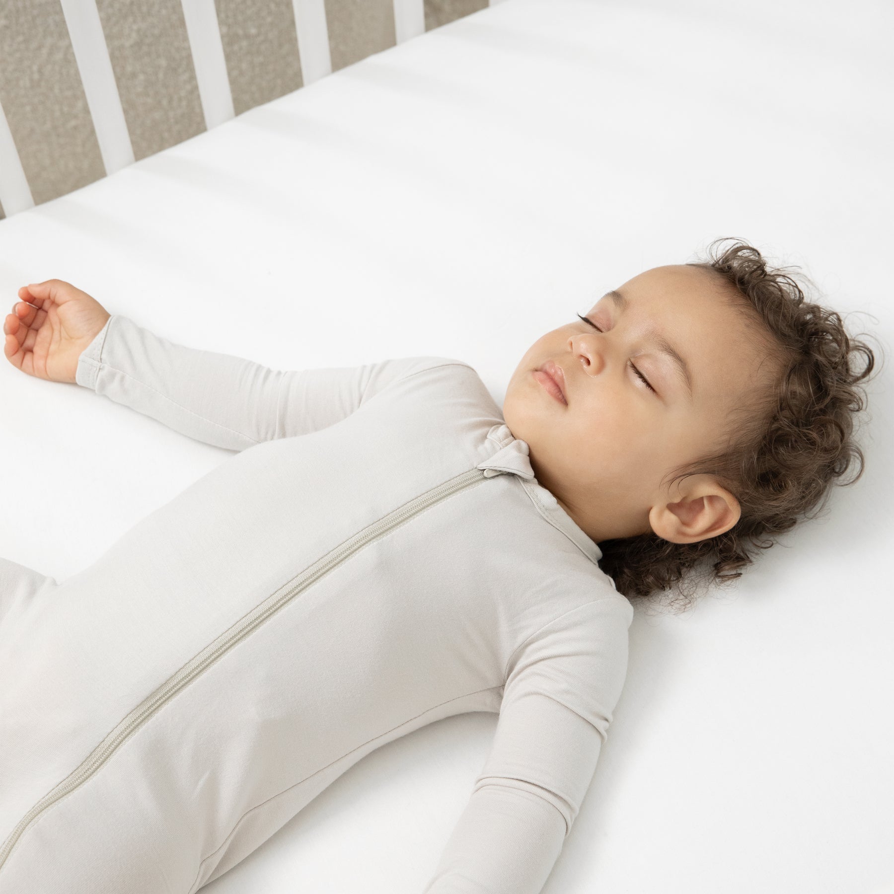 Close up of a sleeping toddler laying in a crib wearing the Zippered Romper in Oat