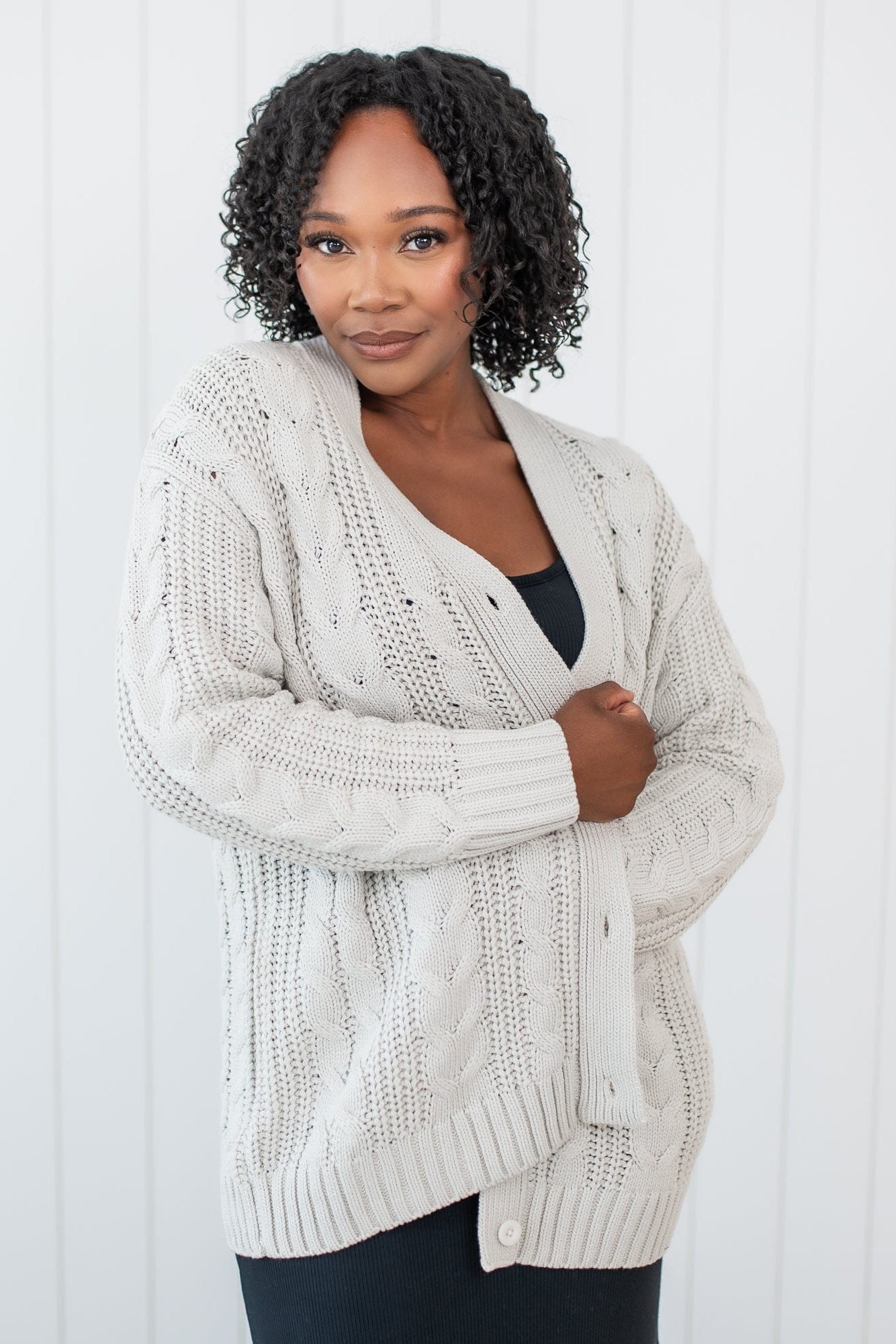 Female standing in front of a white paneled wall wearing the Cable Knit Adult Cardigan in Oat