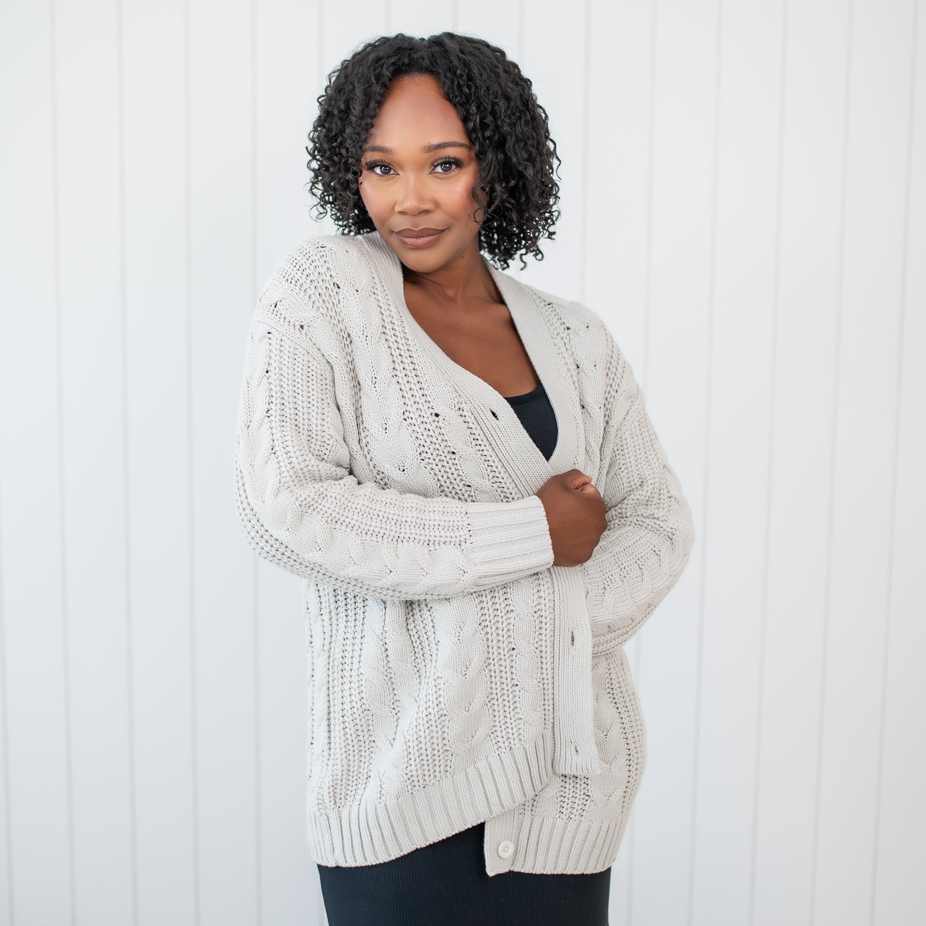 Female standing in front of a white paneled wall wearing the Cable Knit Adult Cardigan in Oat
