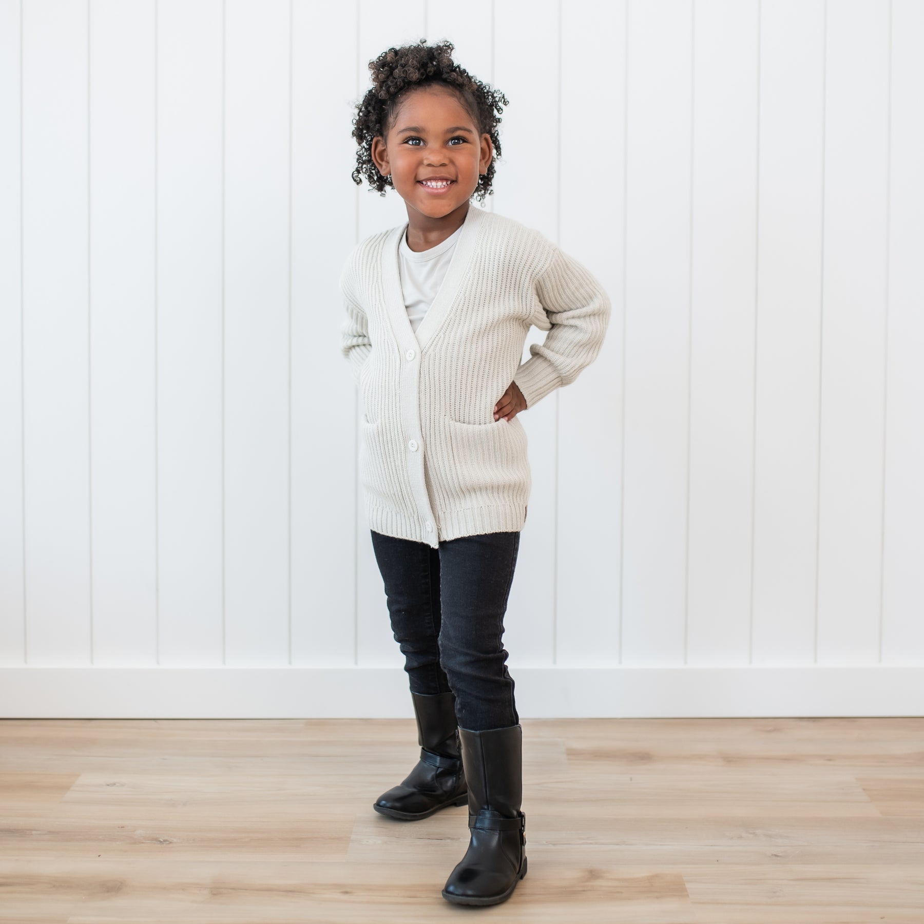 Young girl standing with her hands on her hips wearing the Chunky Knit Oversized Cardigan in Oat with black pants and black boots