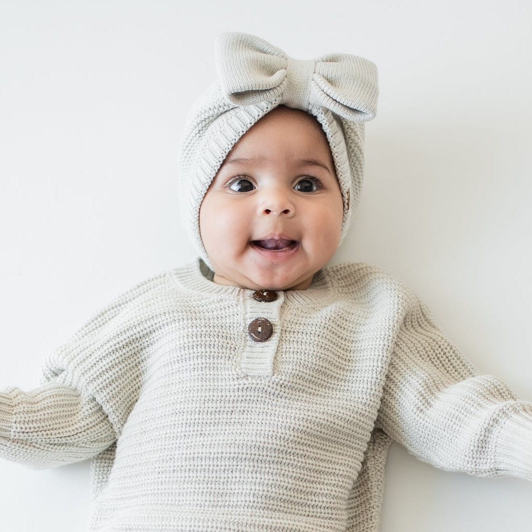 Infant laying down wearing the Chunky Knit Bow Headband in Oat and matching chunky knit romper and knit booties