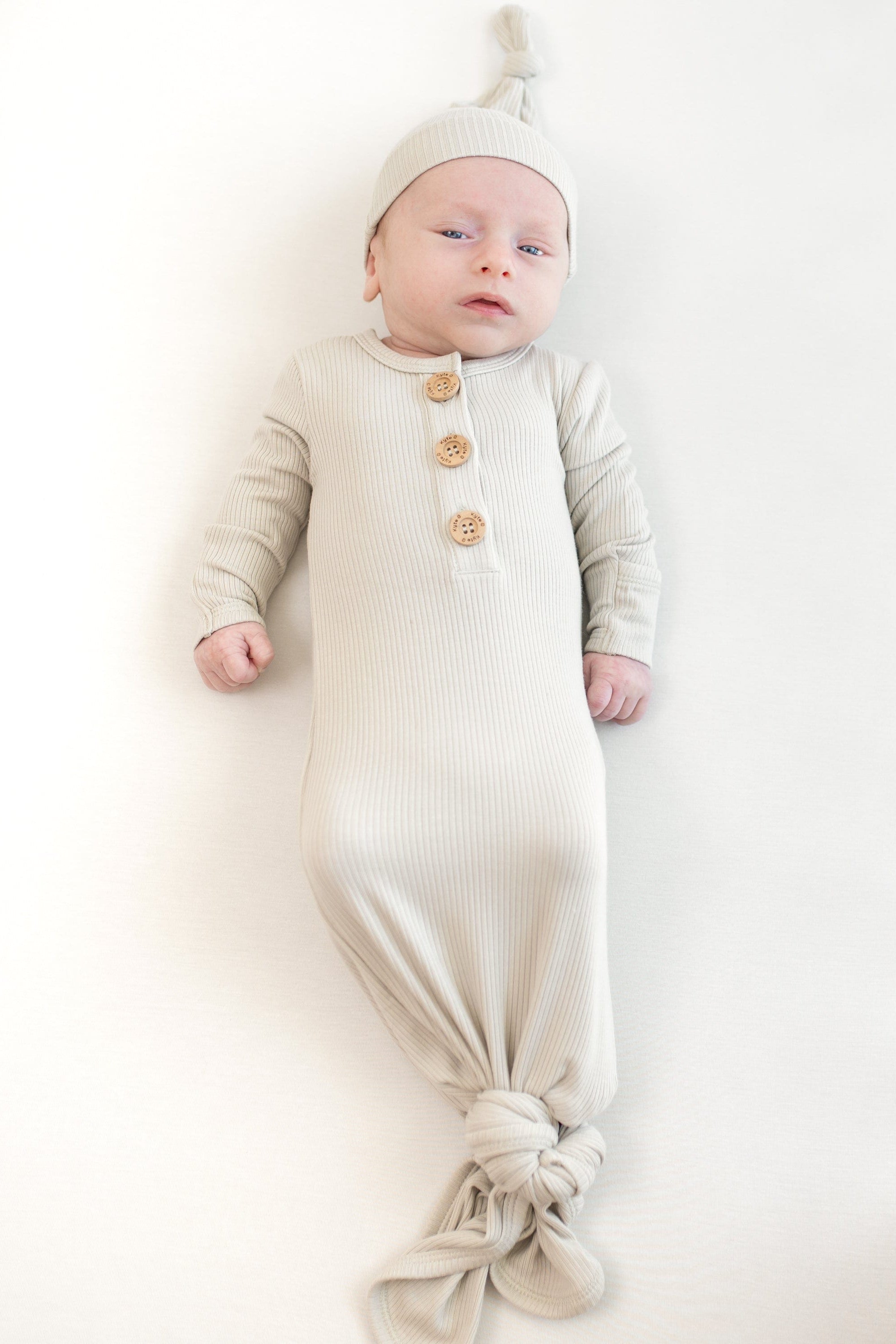 Baby in laying wearing the Kyte Baby Ribbed Knotted Gown with matching hat in the color oat.