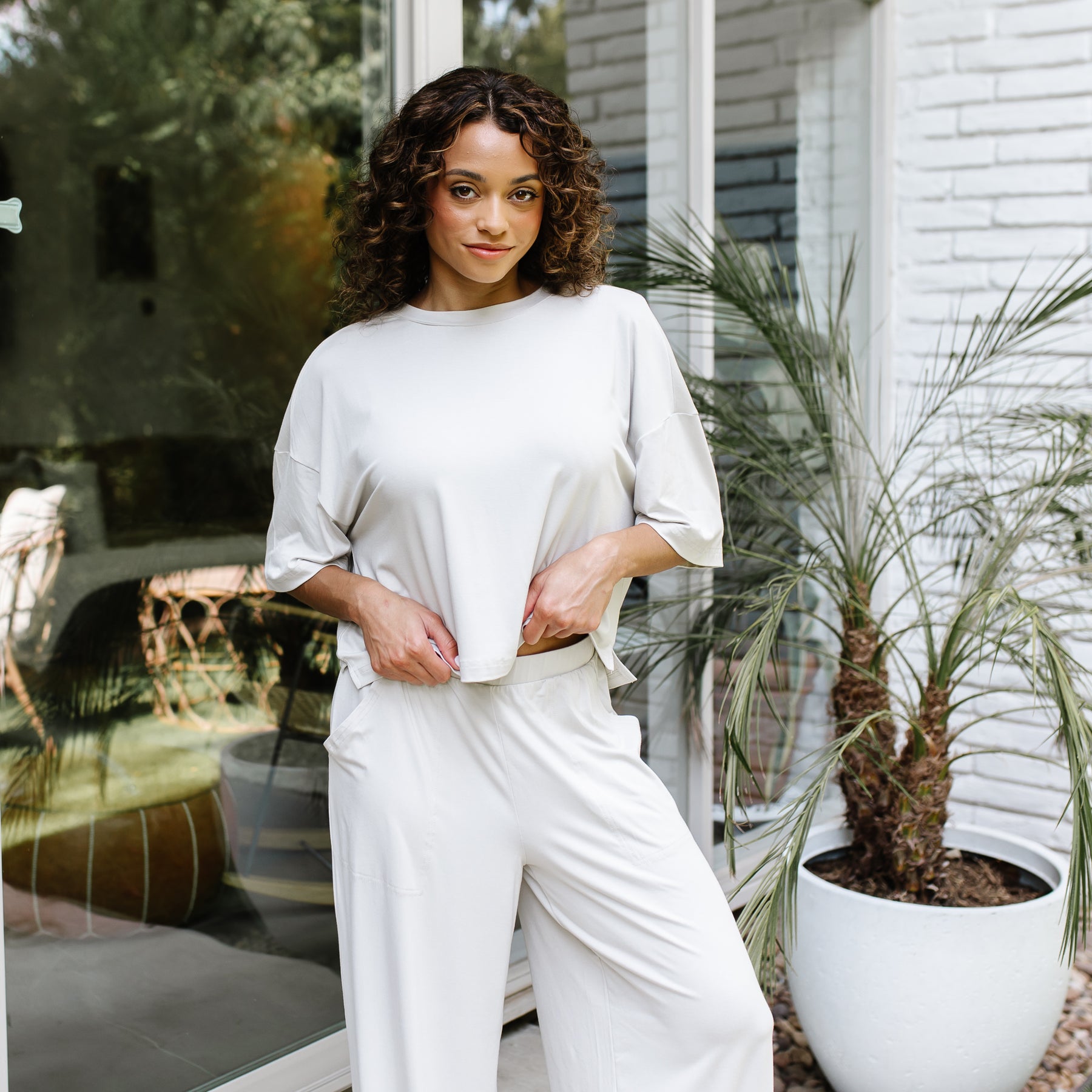 Female model standing in front of a glass window on a white brick house wearing the Women's Short Sleeve Tee in Oat with matching wide leg pants
