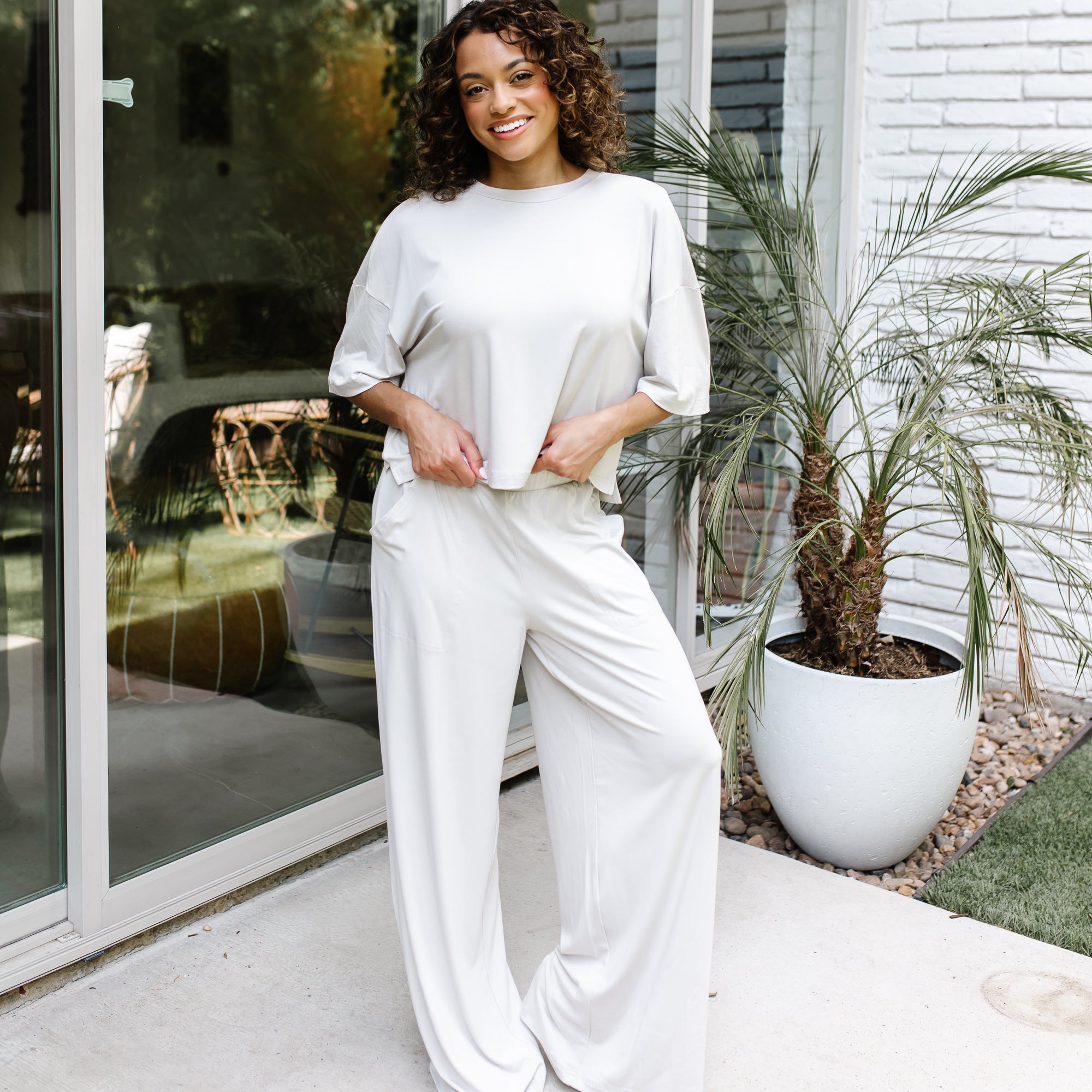 Smiling female standing on a concrete patio in front of glass windows on a white brick house wearing the Women's Wide Leg Pant in Oat and matching women's short sleeve tee