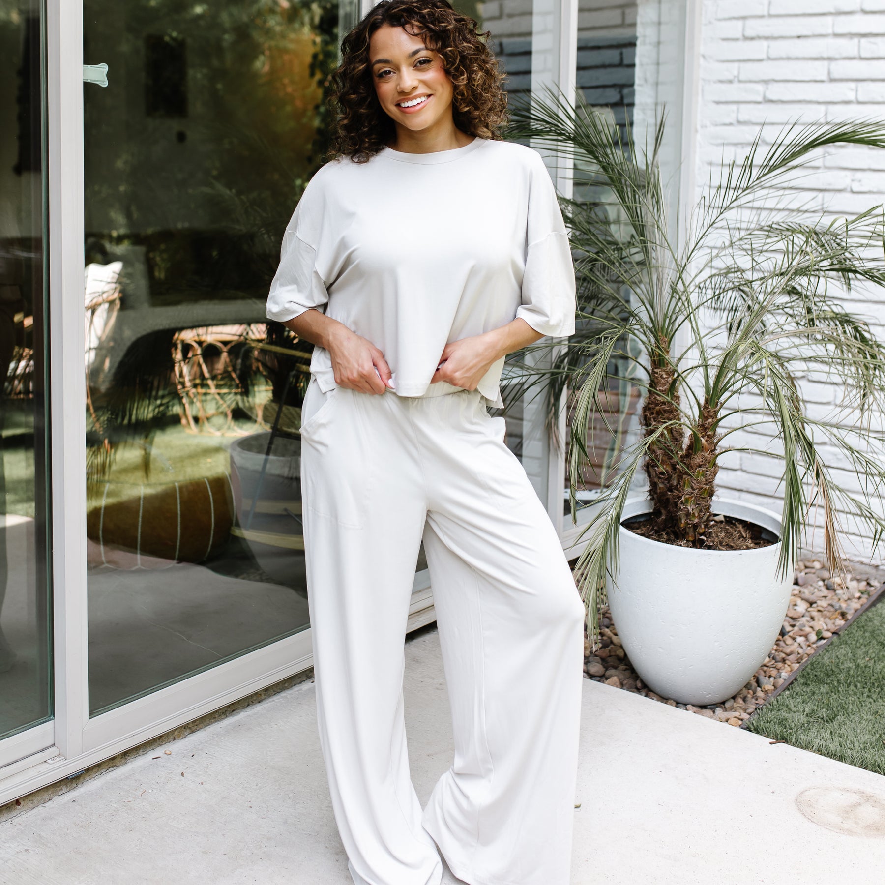 Smiling female standing on a concrete patio in front of glass windows on a white brick house wearing the Women's Wide Leg Pant in Oat and matching women's short sleeve tee