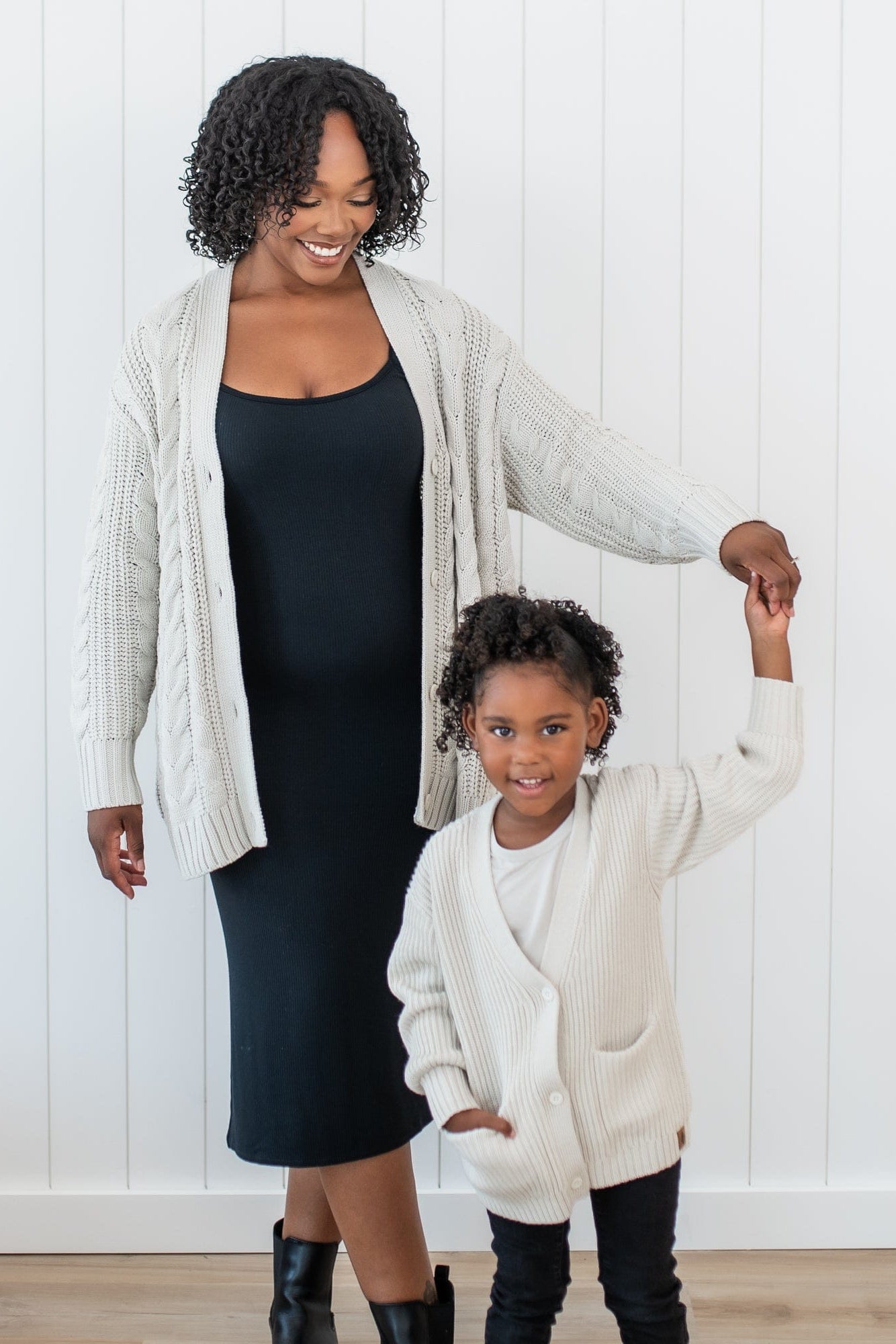 Female standing wearing the Cable Knit Adult Cardigan in Oat holding her daughters hand who is matching in a Chunky knit oversized cardigan