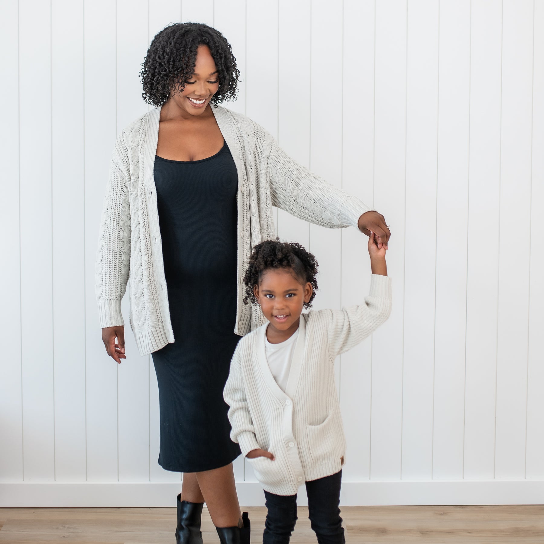 Female standing wearing the Cable Knit Adult Cardigan in Oat holding her daughters hand who is matching in a Chunky knit oversized cardigan