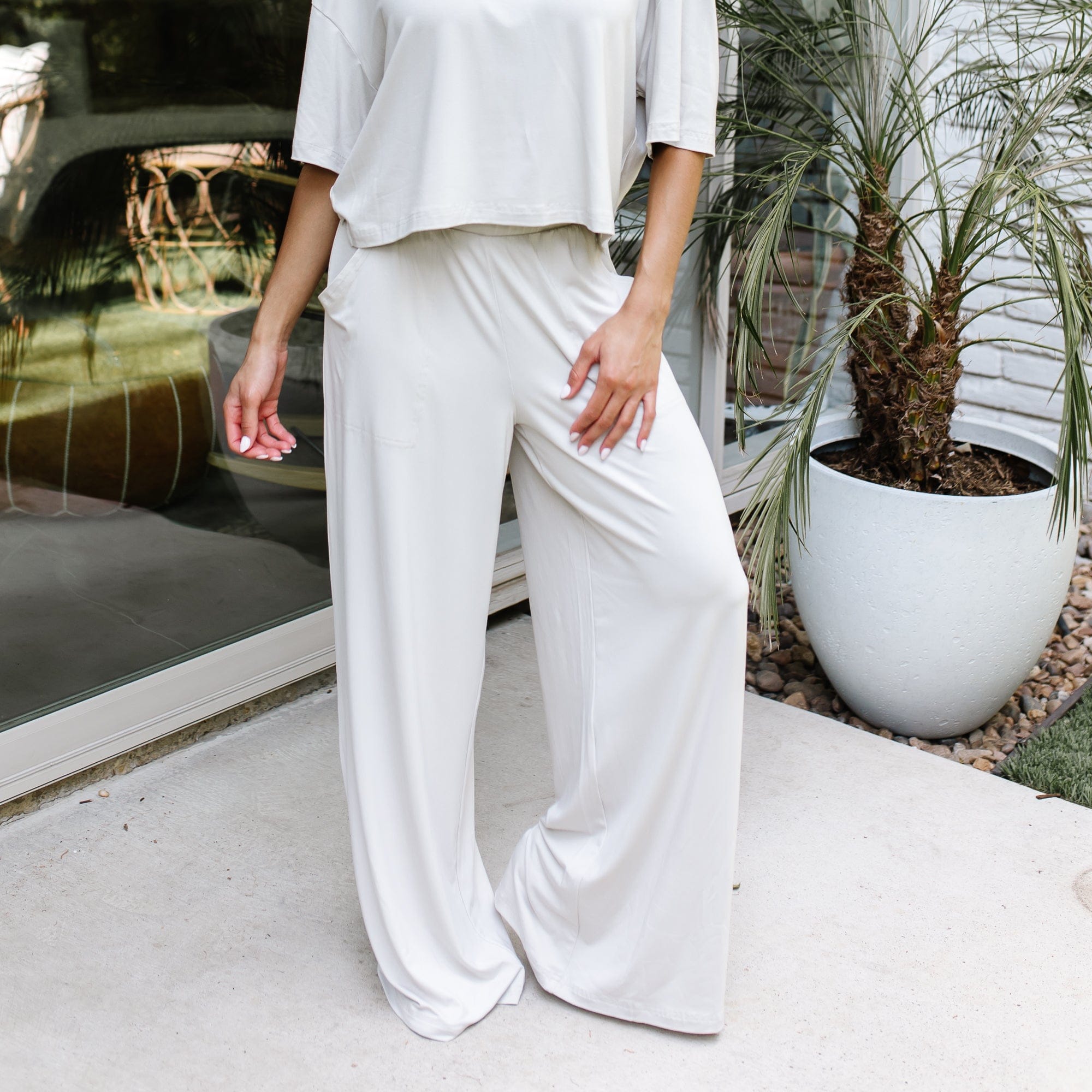 Female model standing on a sidewalk in front of a glass door of a white brick house wearing the Women's Wide Leg Pant in Oat