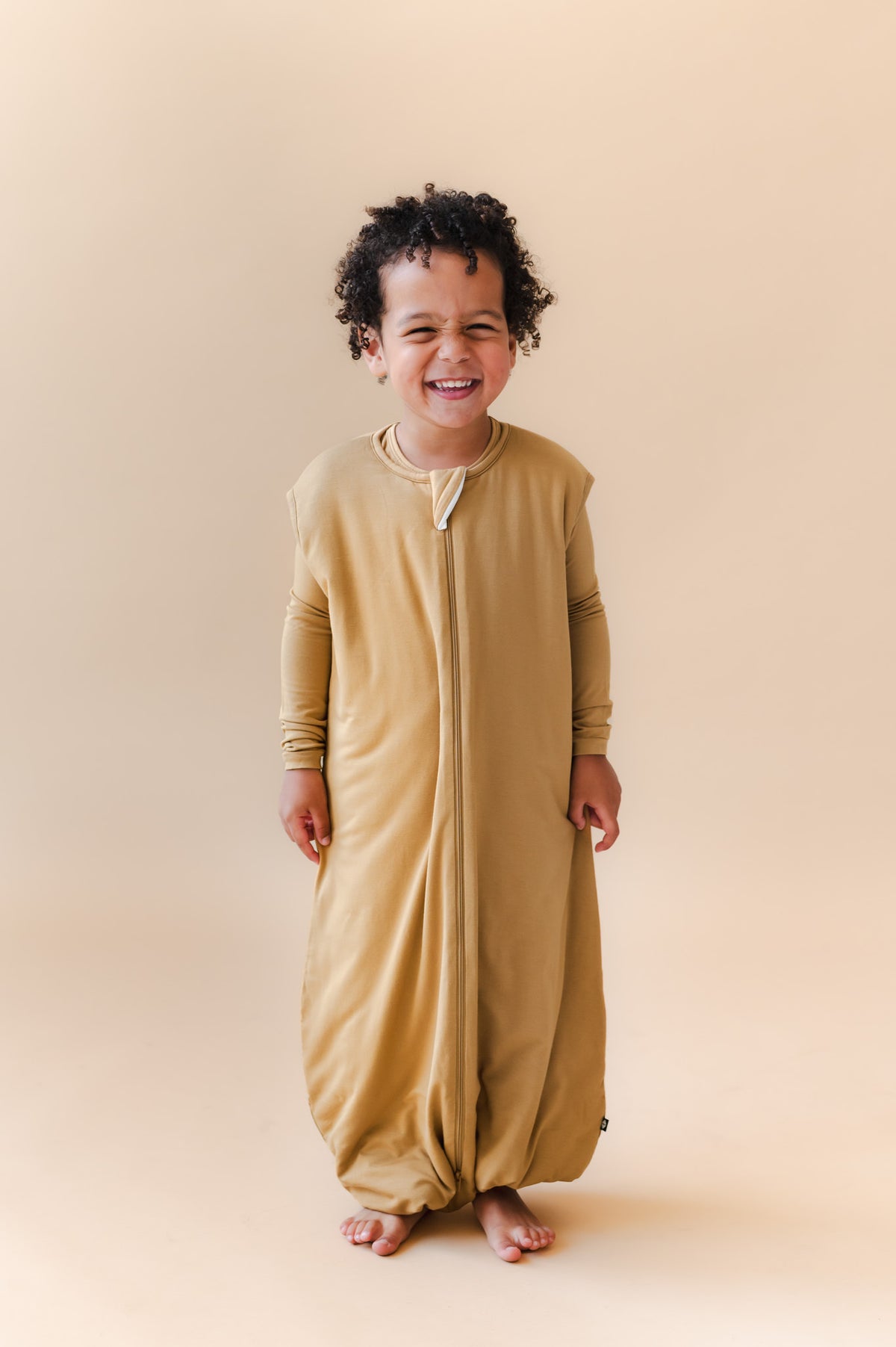 A smiling toddler stands barefoot against a warm beige studio background, wearing a full-length zip-front sleep sack in a mustard yellow color.