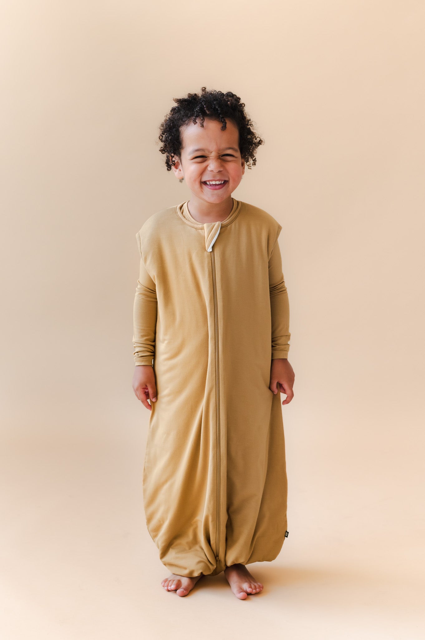 A smiling toddler stands barefoot against a warm beige studio background, wearing a full-length zip-front sleep sack in a mustard yellow color.