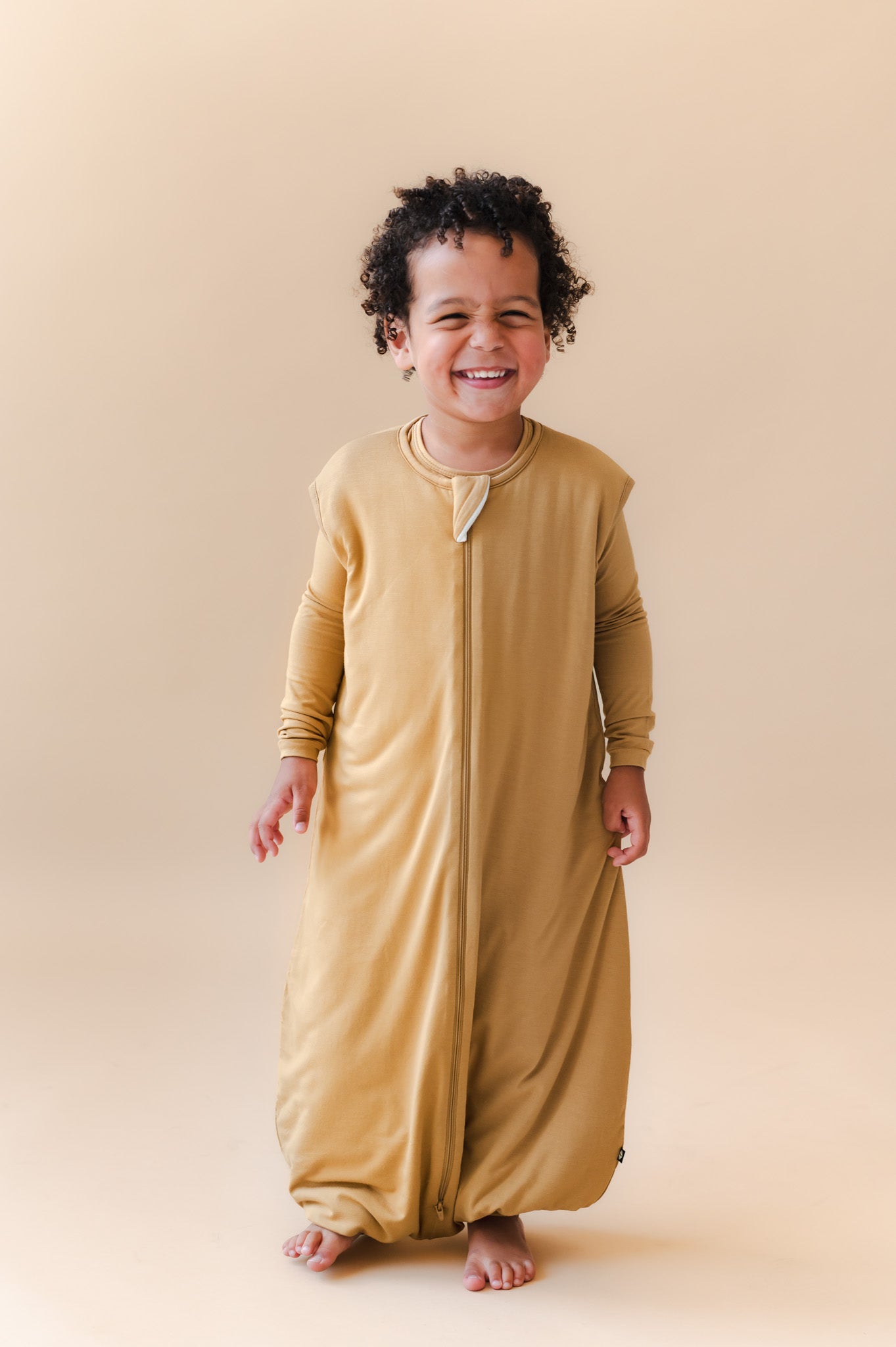 A smiling toddler wearing a mustard yellow full-length zip-up sleep sack walker, standing against a solid beige studio background.