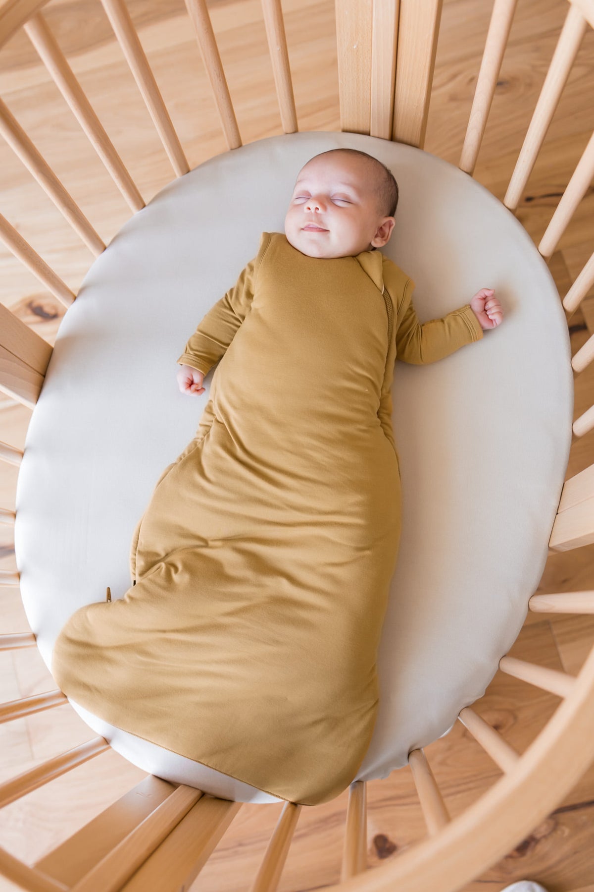 A newborn baby wearing a mustard yellow long-sleeve sleep sack, lying on a white mattress inside a wooden crib.