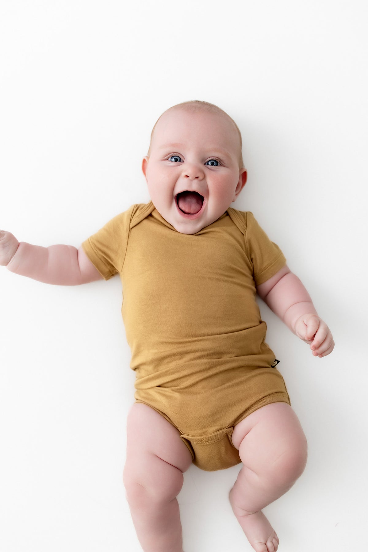 A smiling baby lying on their back wearing a mustard yellow short-sleeve bodysuit against a clean white background.