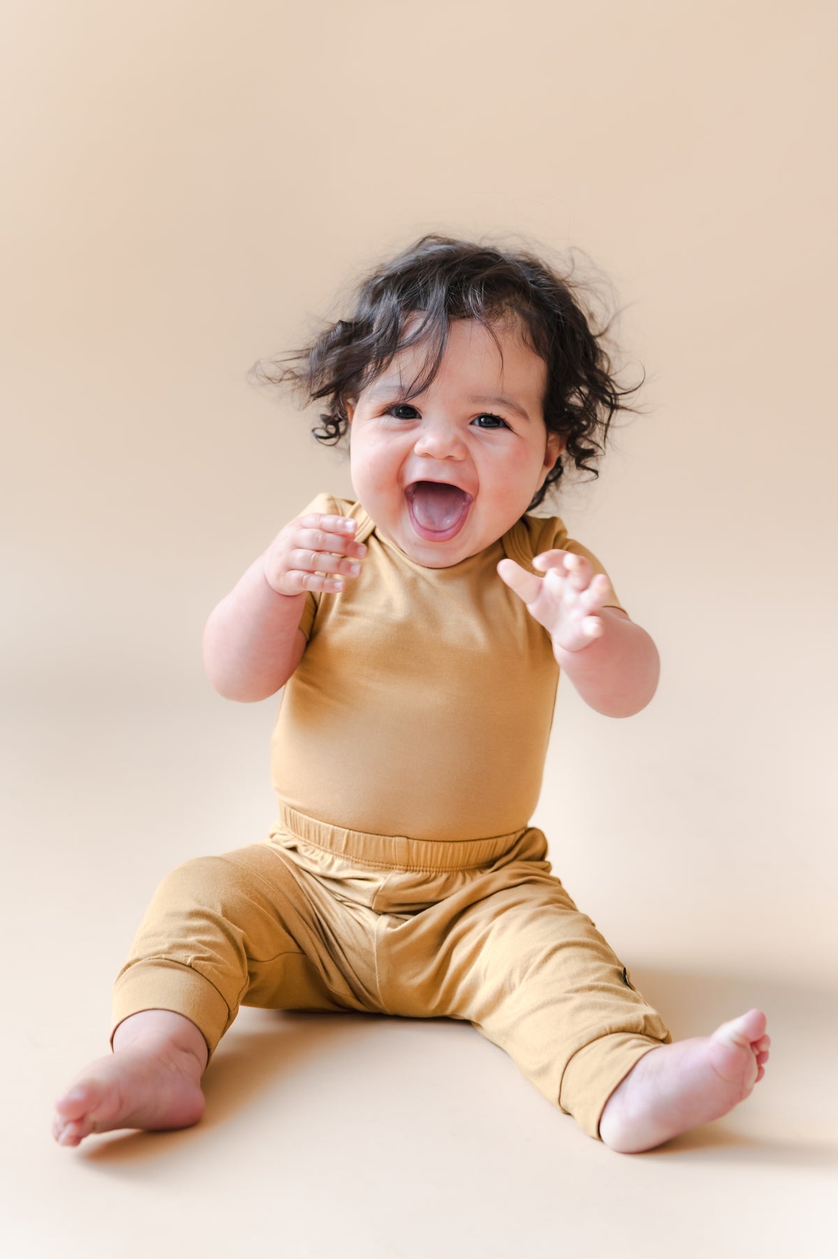 Baby smiling in Pant in Ochre over similar color bodysuit on a beige background