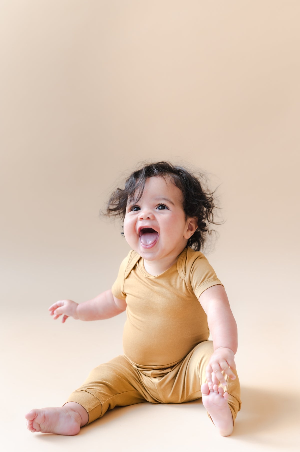 Baby laughing in Pant in Ochre over similar color bodysuit on a beige background