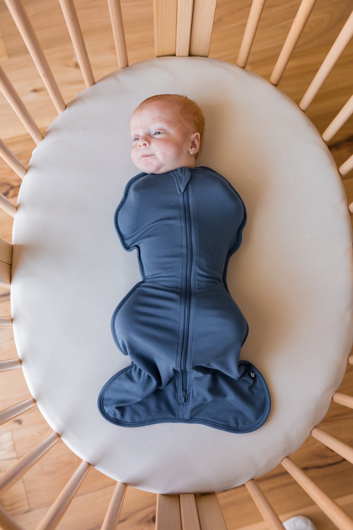 A baby lying on their back inside a wooden crib, swaddled in a dark slate blue zippered swaddle bag. The baby is resting on a round cream-colored mattress pad.