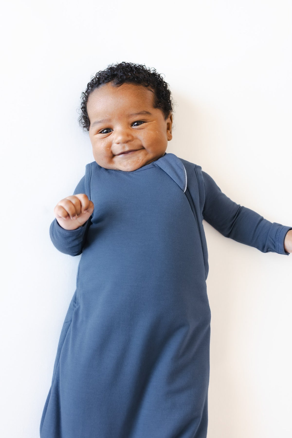 A smiling baby wearing a slate blue sleep sack, posed against a clean white background.
