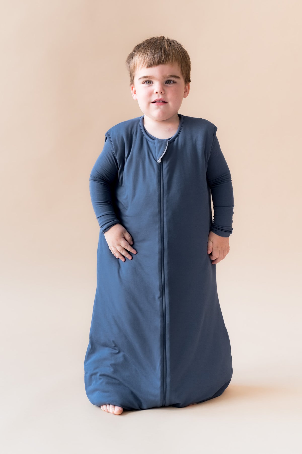 A toddler wearing a navy blue zippered sleep sack standing against a solid beige studio background.