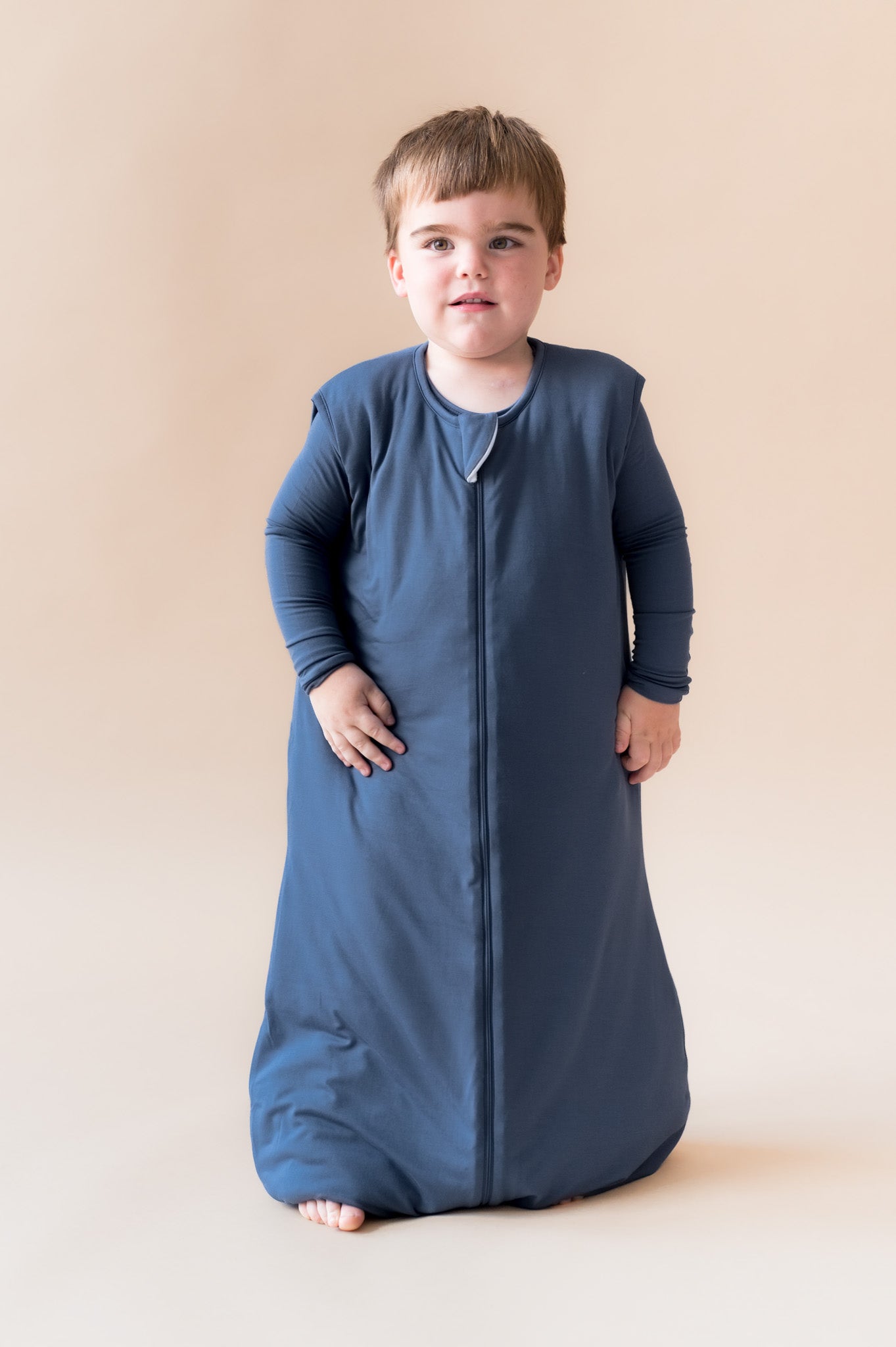 A toddler wearing a navy blue zippered sleep sack standing against a solid beige studio background.