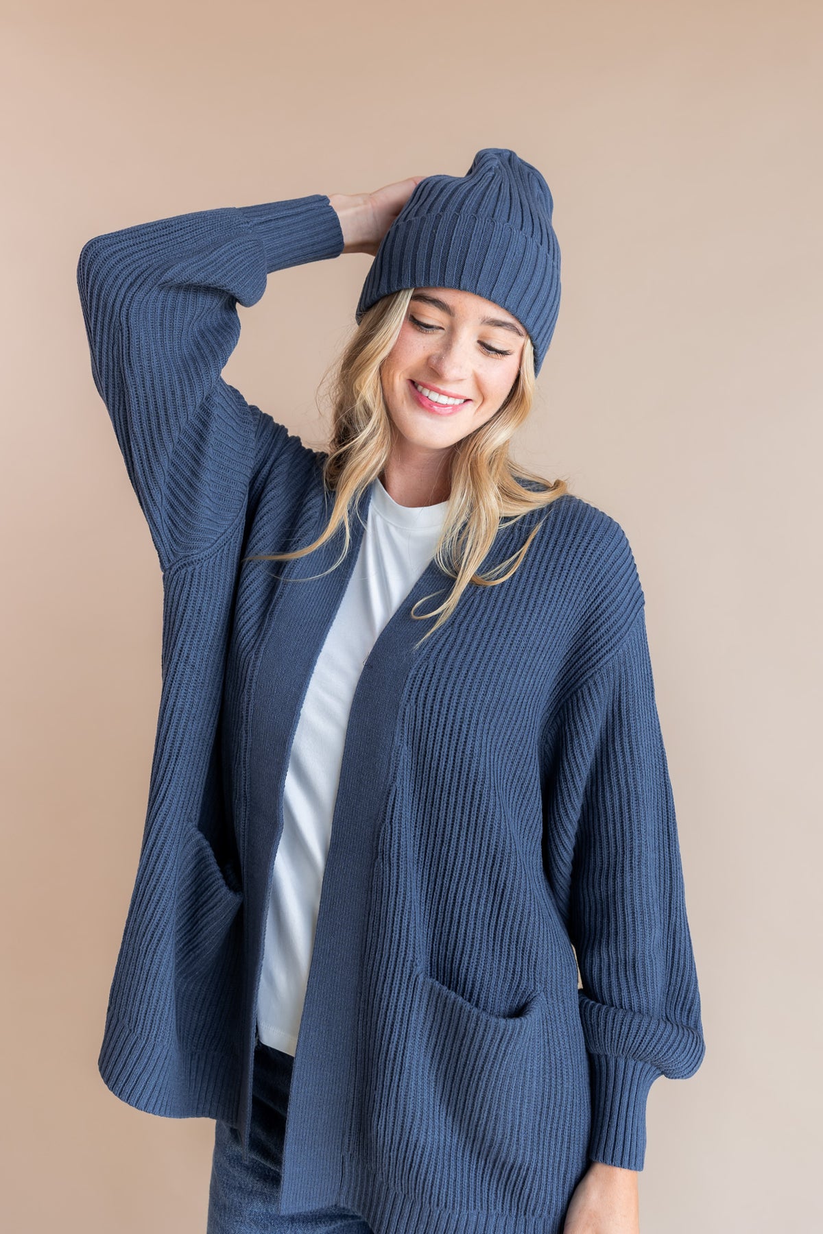 Modal posing in Chunky Knit Adult Ribbed Beanie in Oxford over white T-shirt, blue jeans and Chunky knit cardigan in oxford on a beige background