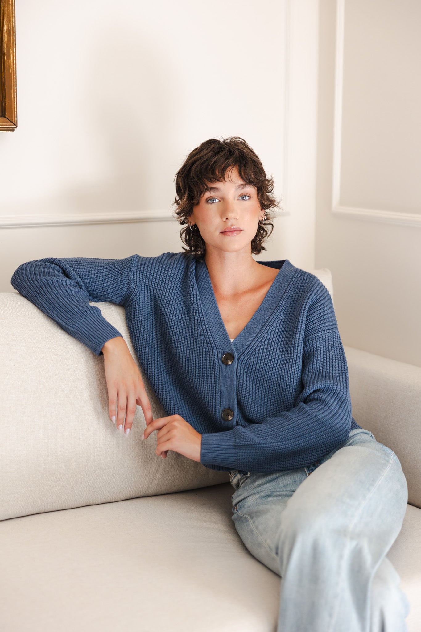 Modal sitting on a white couch wearing Chunky Knit Women's Cropped Cardigan In Oxford over light blue jeans
