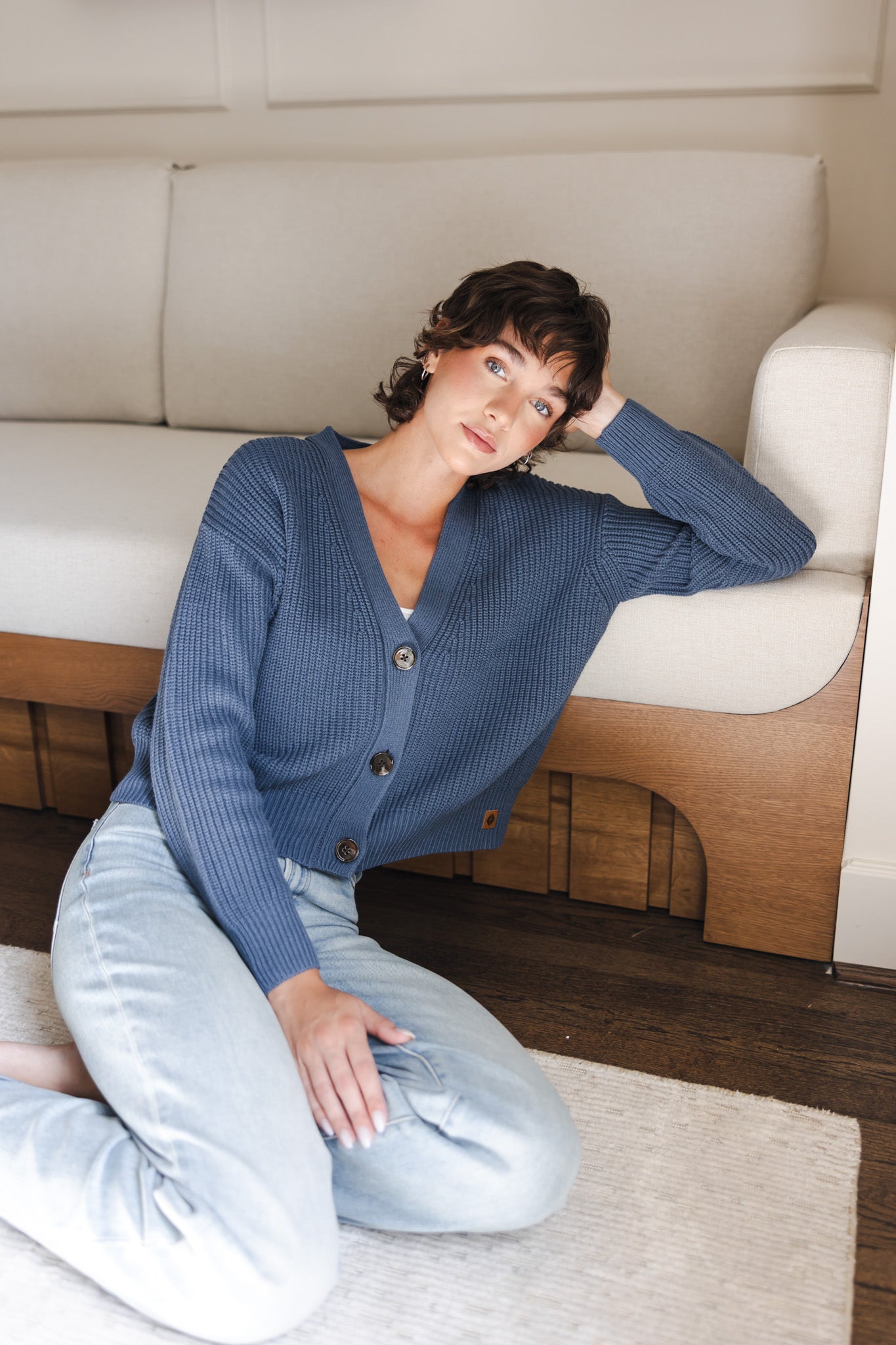 Modal sitting against a white sofa wearing Chunky Knit Women's Cropped Cardigan In Oxford over light blue jeans