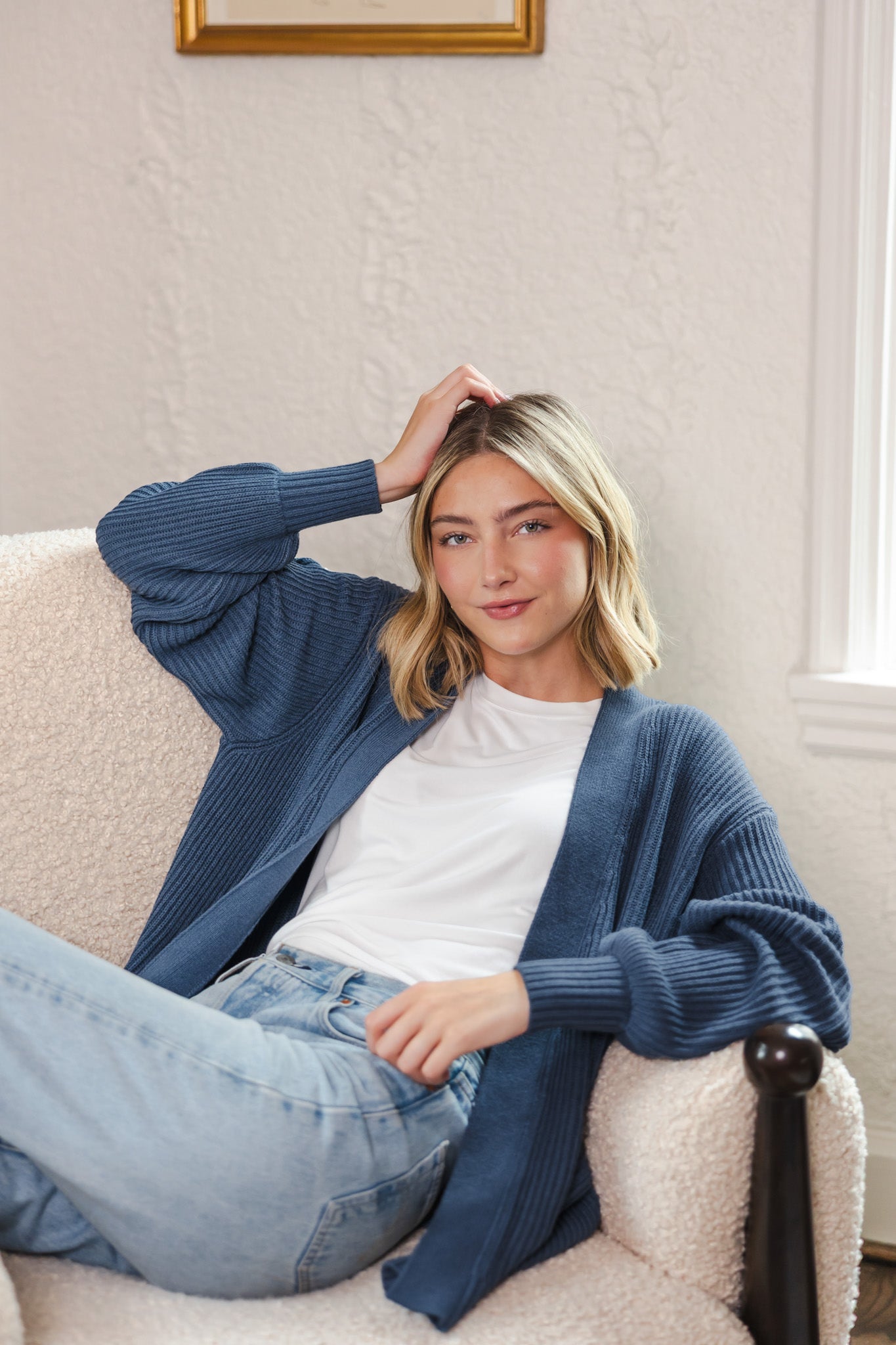 Modal sitting on white sofa wearing chunky knit oversized cardigan in oxford over blue jeans and white t-shirt 
