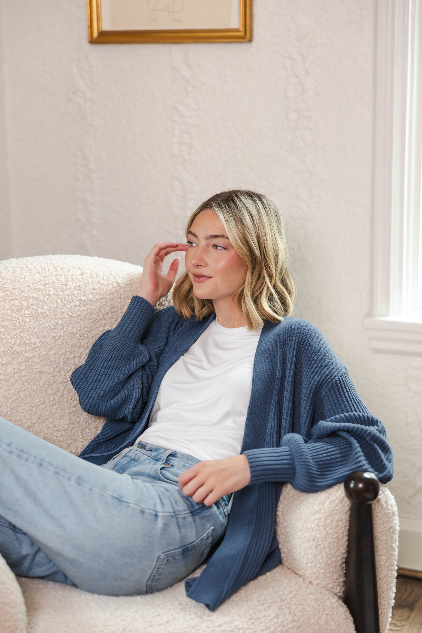 Modal sitting on white sofa wearing chunky knit oversized cardigan in oxford over blue jeans and white t-shirt 
