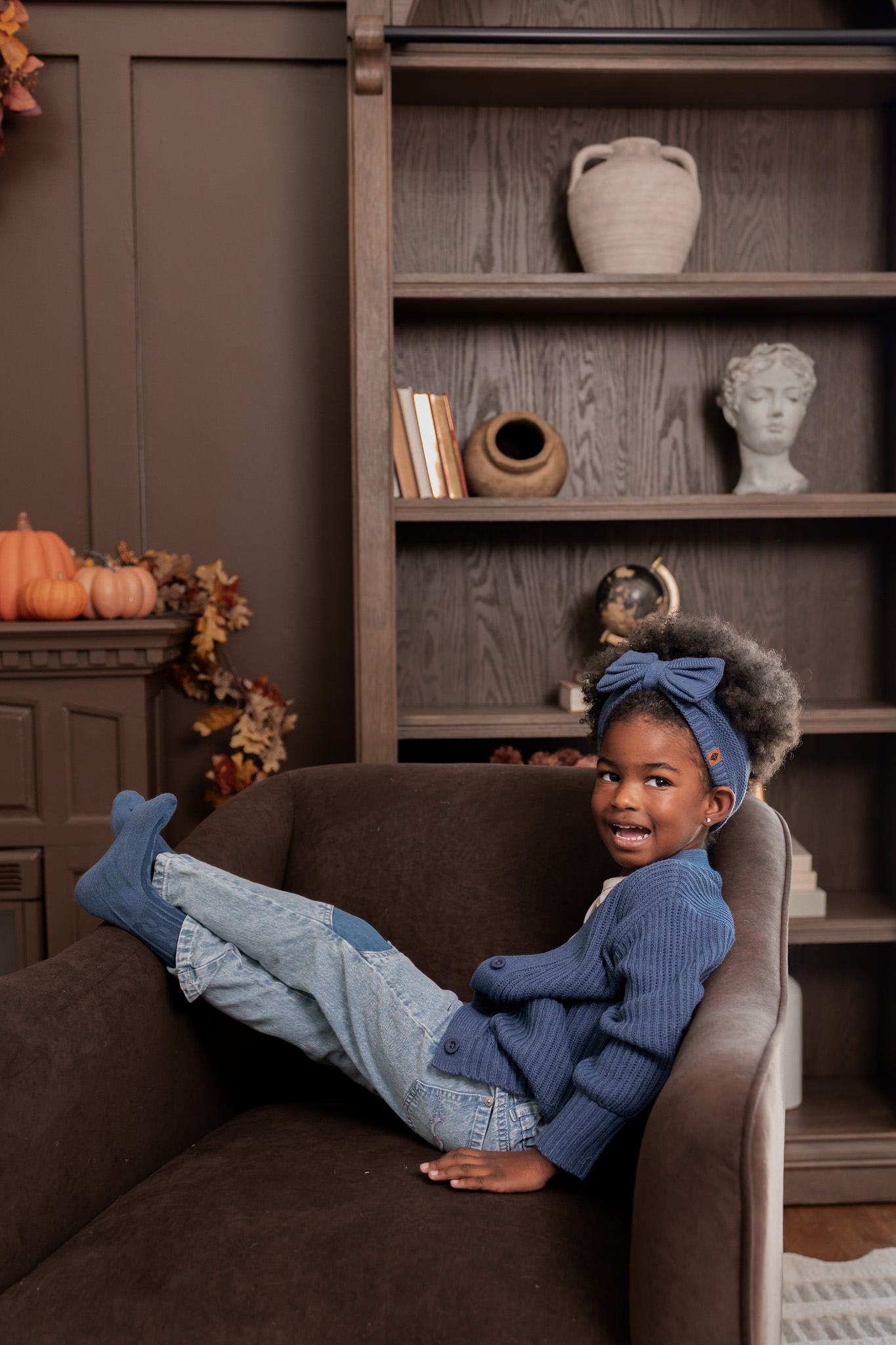 Girl lying on brown sofa wearing Chunky Knit Oversized Cardigan in Oxford over White T-shirt, Chunky knit bow headband, blue jeans and socks in oxford