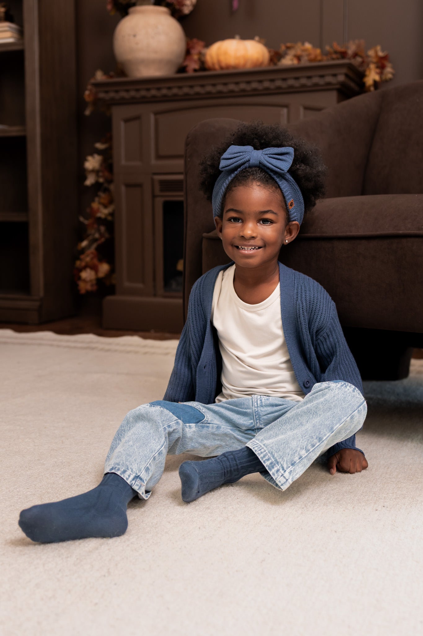Girl sitting on a white carpet wearing Chunky Knit Oversized Cardigan in Oxford over White T-shirt, blue jeans,  Chunky knit bow headband and socks in oxford