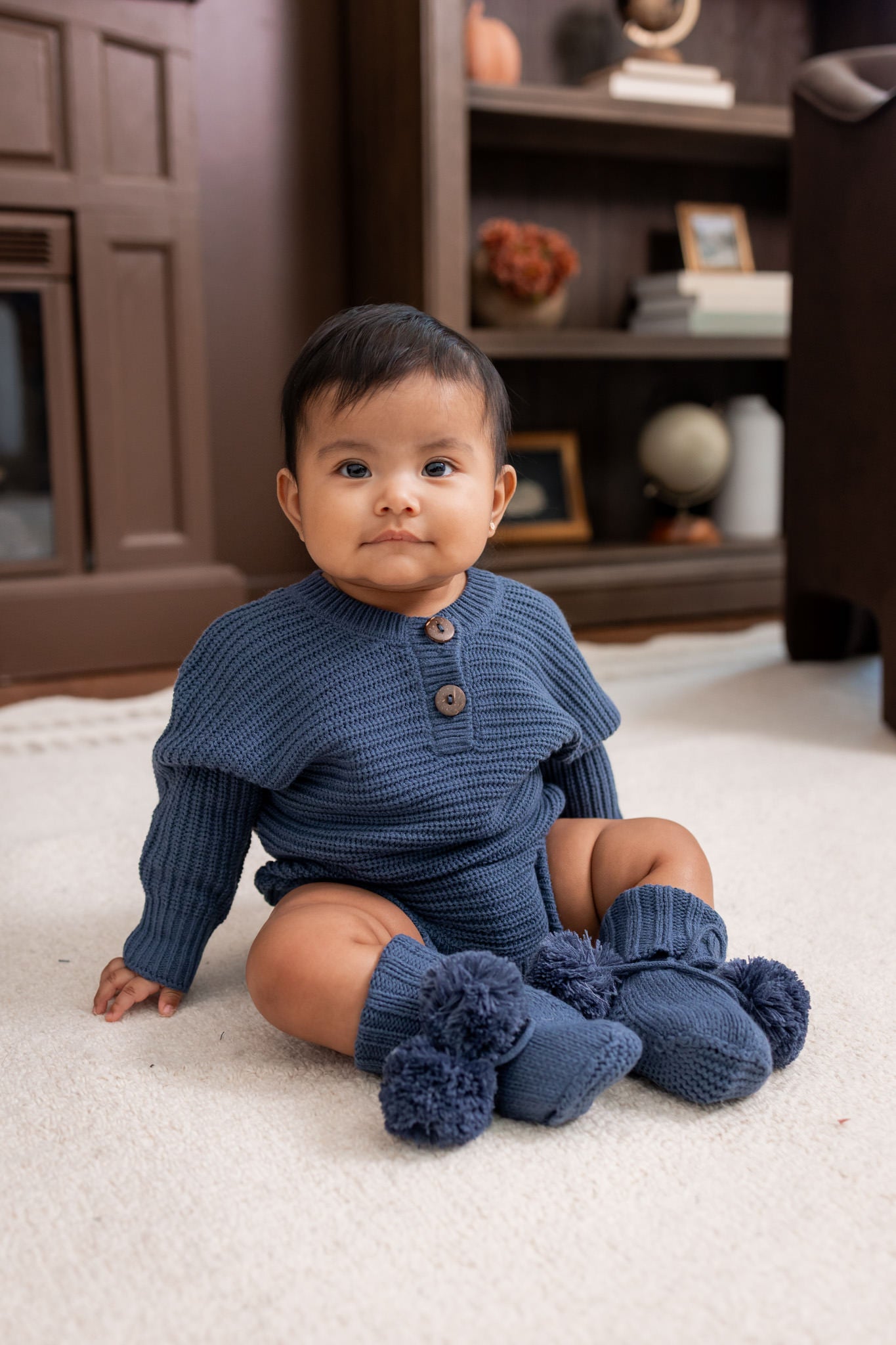 Baby sitting on a white carpet wearing Chunky Knit Romper in Oxford and Knit booties in oxford