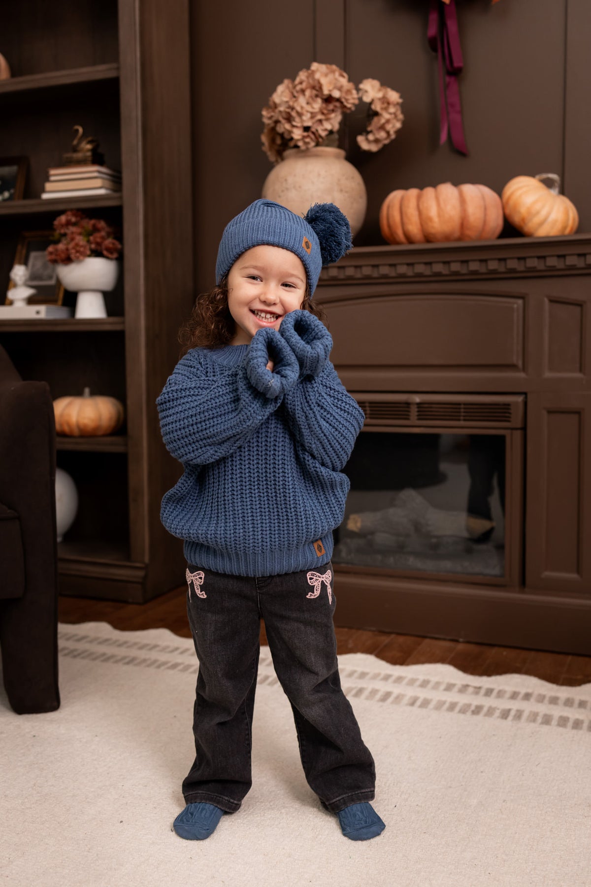 A young child wearing a cozy blue knit beanie with a pom-pom, a chunky blue sweater, and dark pants, standing in front of a fireplace in a decorated living room.