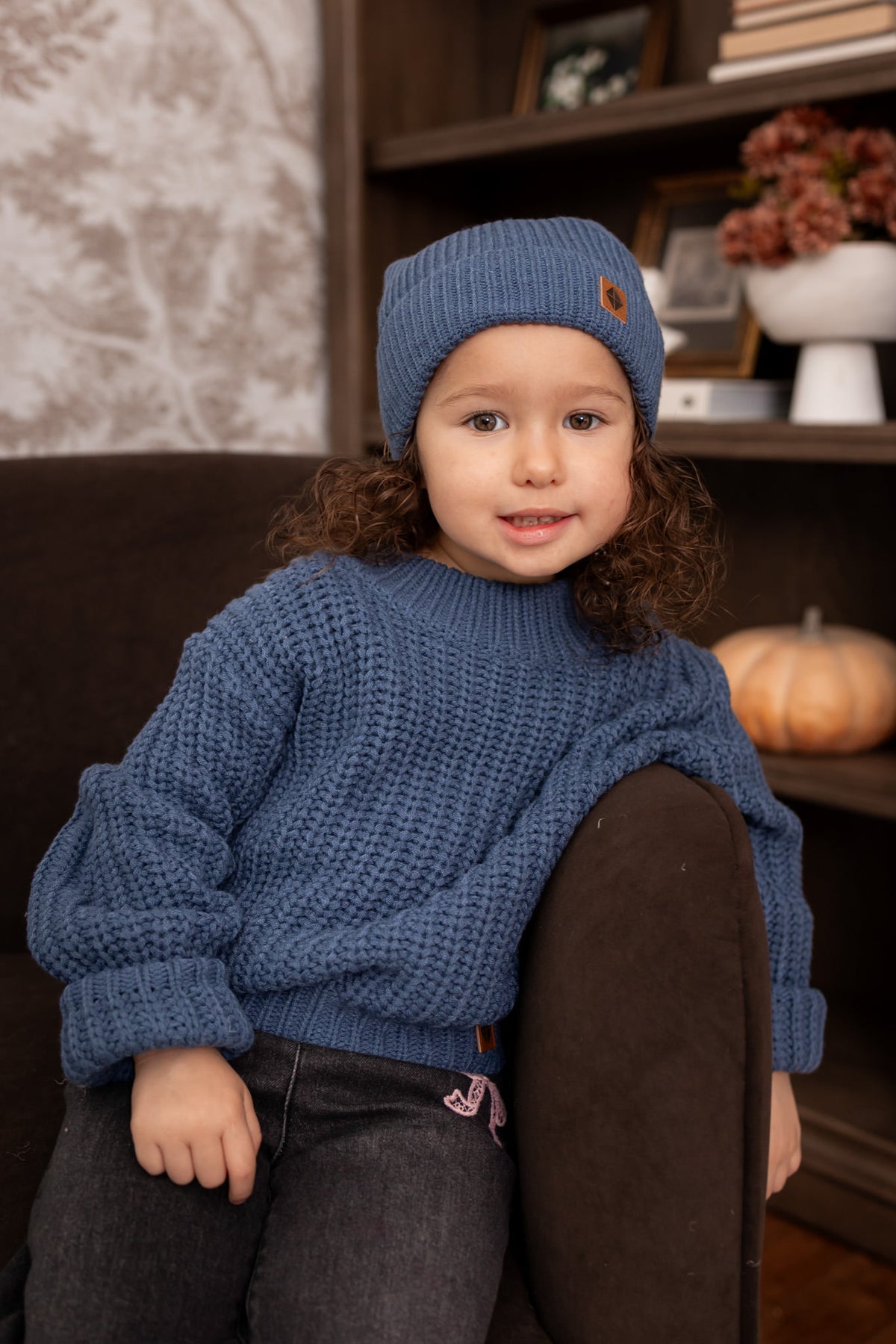 Girl sitting on brown sofa while wearing Chunky Knit Sweater and chunky knit pom beanie in Oxford over black pants 