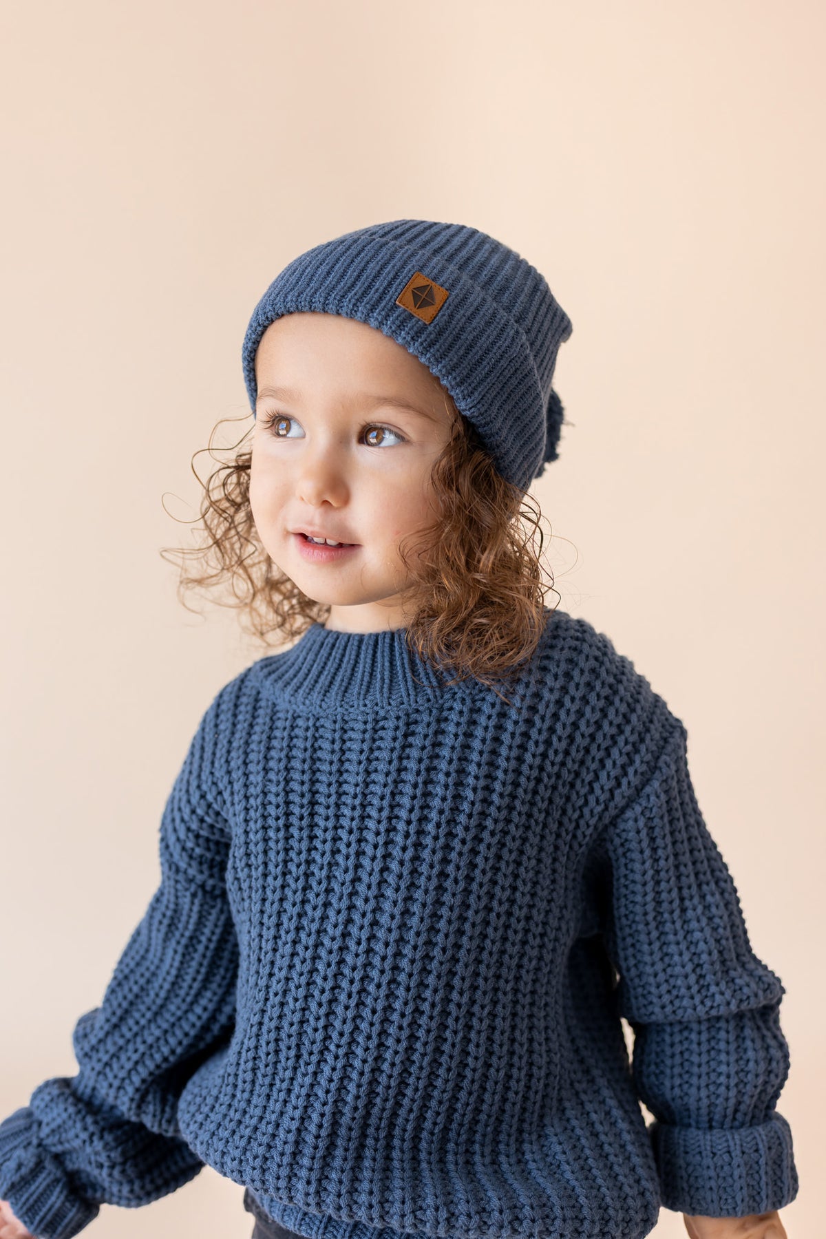 A toddler with curly light brown hair wearing a chunky navy blue knit beanie with a small brown logo patch and a matching navy blue ribbed knit sweater, posed against a solid light beige background.
